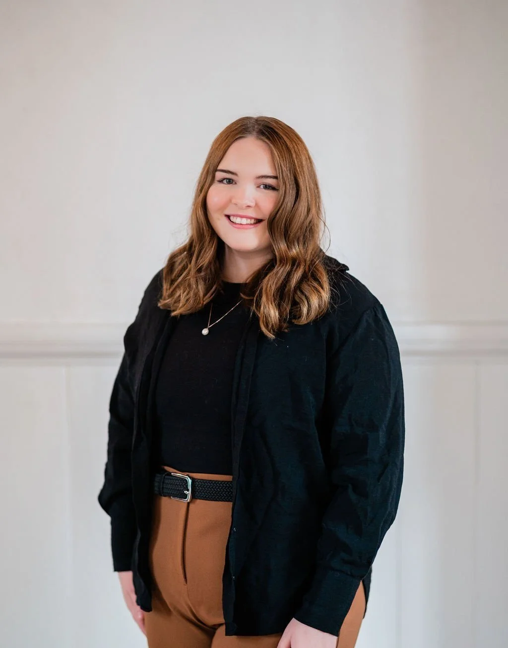 Smiling young woman with wavy brown hair wearing a black jacket, black top, and tan pants, standing indoors against a plain off-white wall.