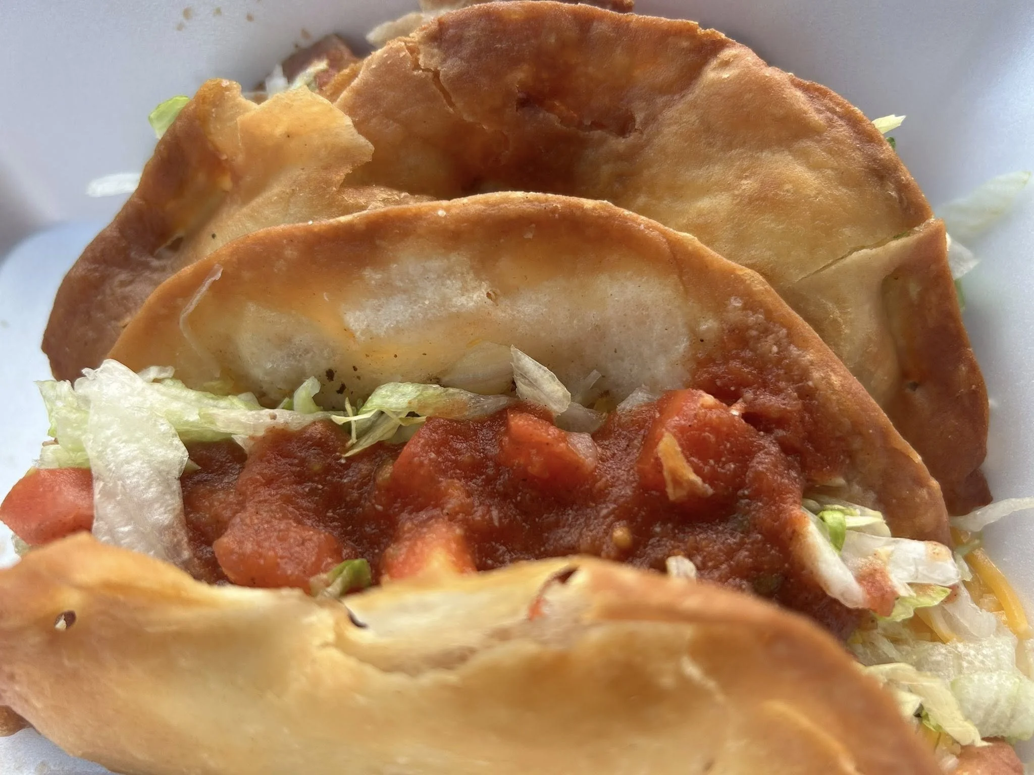 Three tacos with fried shells, shredded lettuce, grated cheese, and tomato-based salsa.