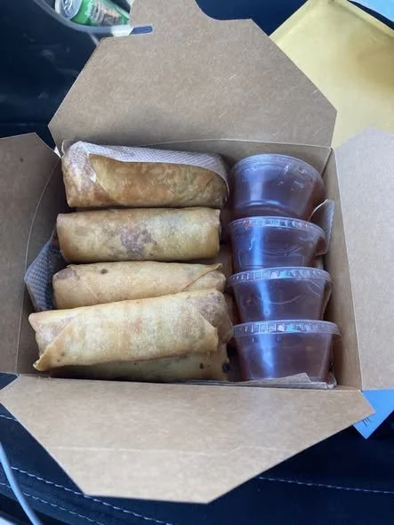 Six fried spring rolls with four containers of dipping sauce in a takeout box.