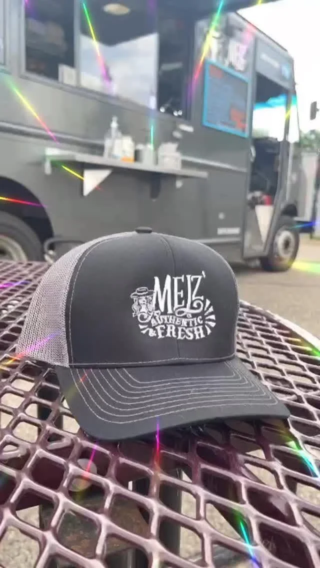 A black and gray truck with a menu or advertisement on its side, parked outdoors. In the foreground, a black trucker cap with white text reading 'MEZ Authentic & Fresh' is placed on a metal picnic table.