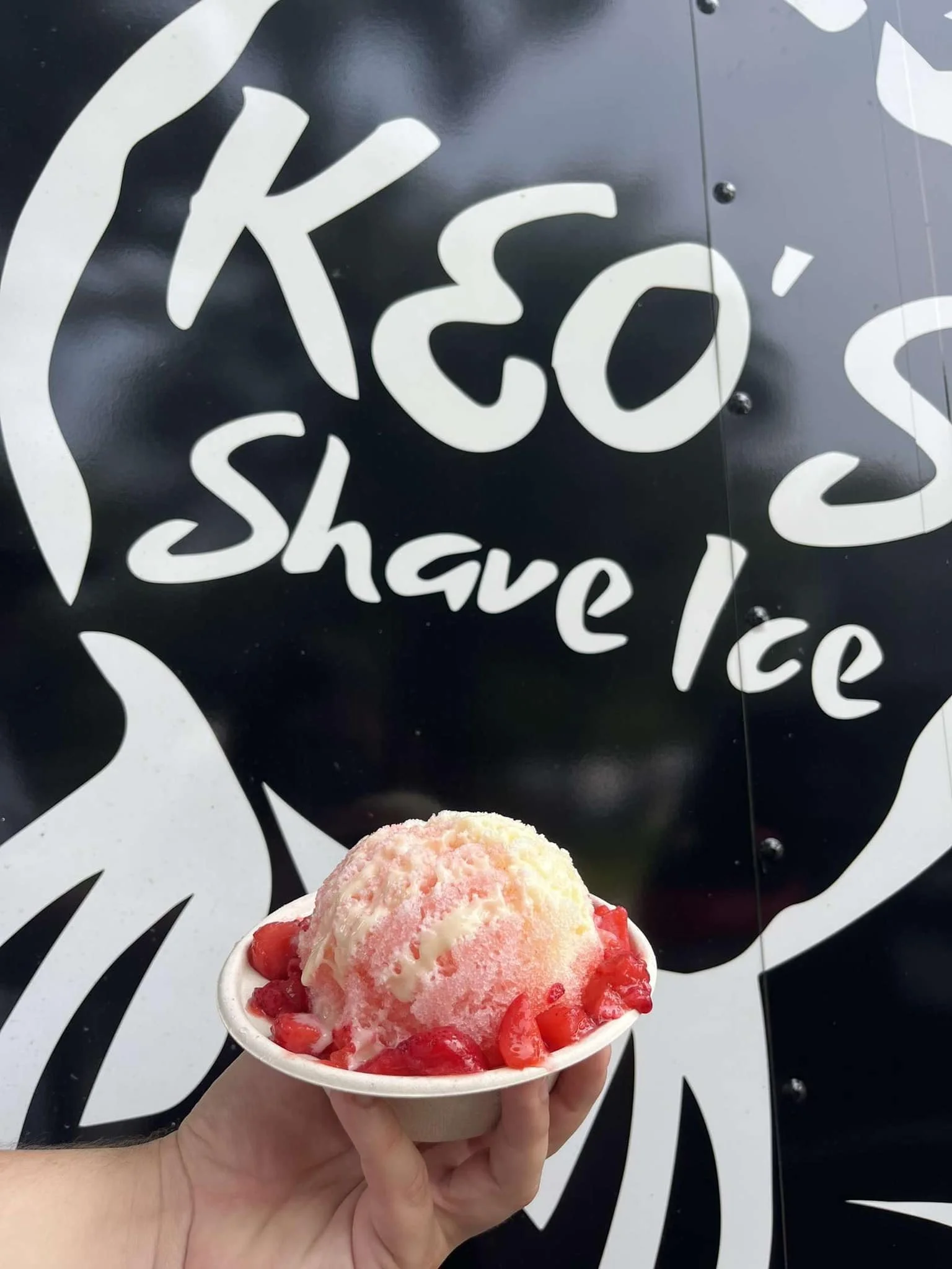 A hand holding a cup of strawberry ice cream topped with strawberries, in front of a black sign with white text that reads 'KCO' and 'shake ice'.