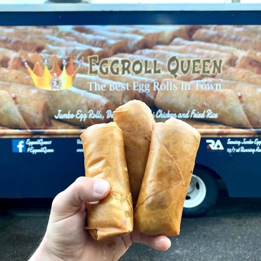 A person's hand holding three egg rolls in front of a food truck that reads 'Eggroll Queen The Best Egg Rolls in Town'.
