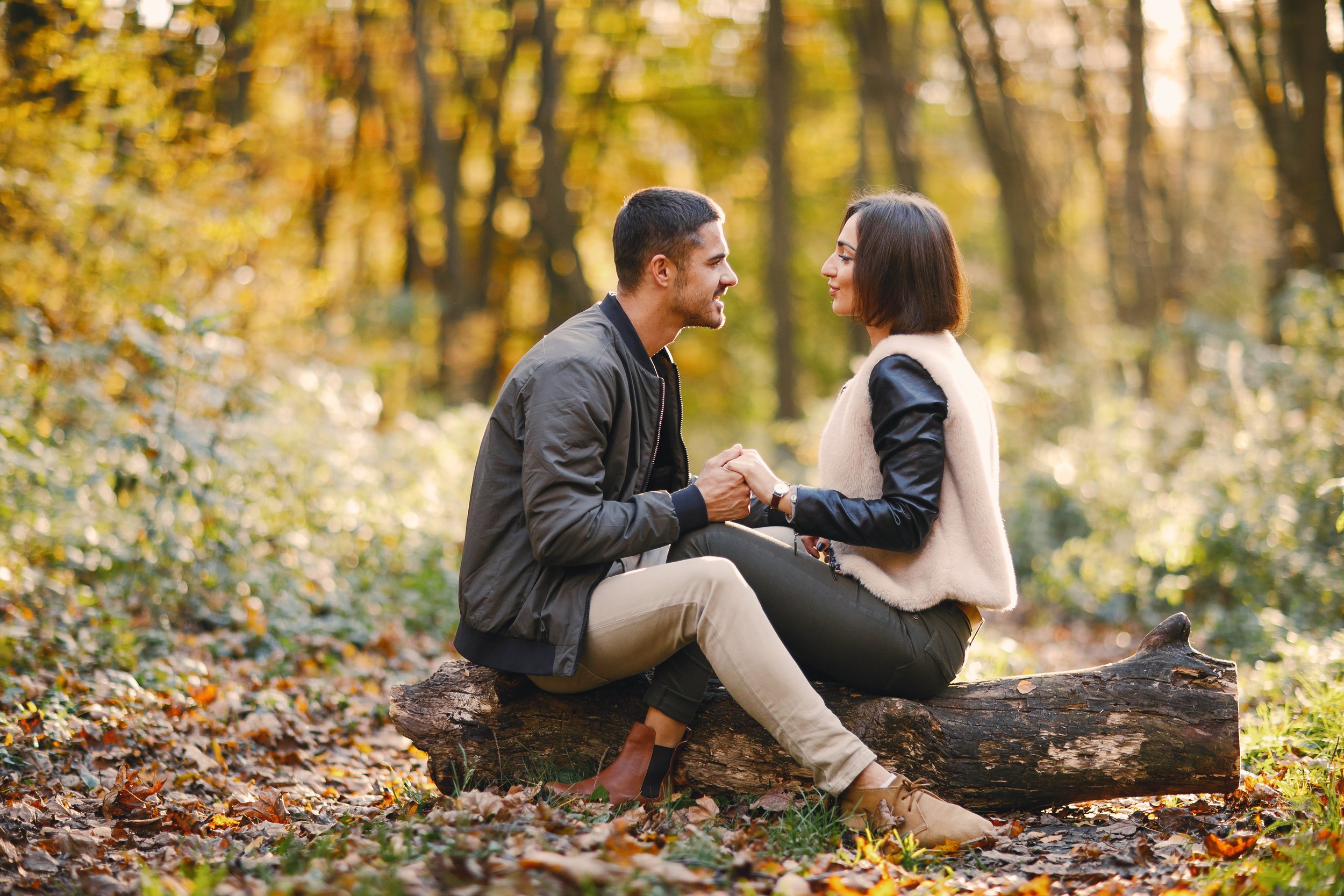 Photo of a young couple sitting on a log facing each other and smiling while holding hands in an autumnal forest