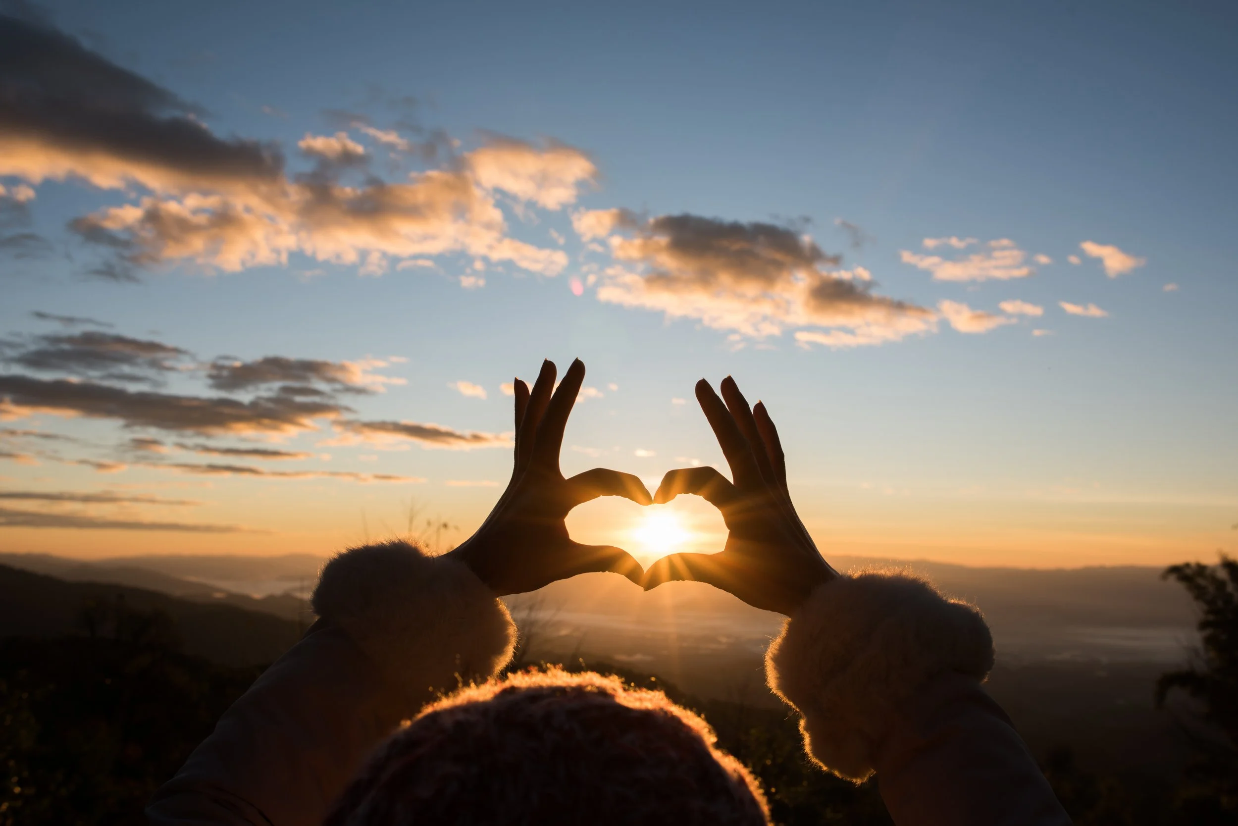 Photo showing the silhouette of a person making a heart shape using their hands over the setting sun