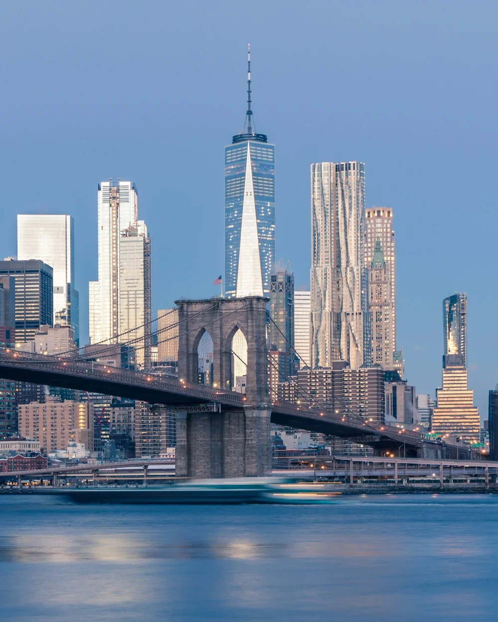 Skyline of Manhattan, New York City, featuring the Brooklyn Bridge in the foreground and One World Trade Center in the background during twilight.