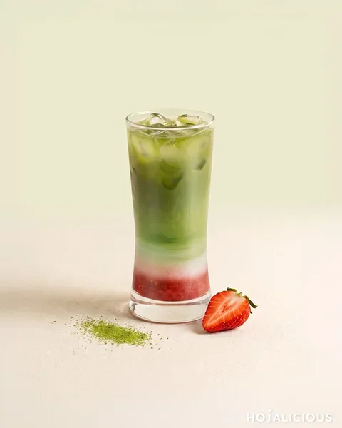 A strawberry matcha recipe layered with strawberry jam, fresh milk, Jin Xuan tea and matcha in a tall clear glass, with a fresh strawberry and matcha powder beside it.