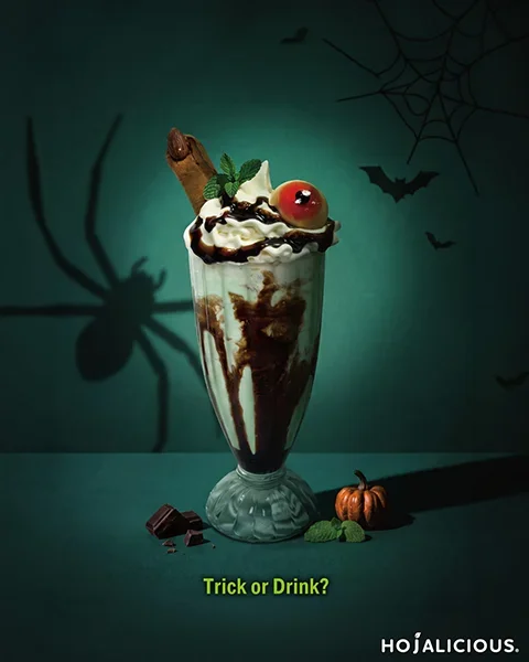 Witch’s Mint Chocolate Milkshake served in a clear tall glass with a pale green shake, dark chocolate drips, whipped cream, chocolate sauce, a witch finger cookie, an eyeball gummy, and a mint leaf for a spooky Halloween presentation.