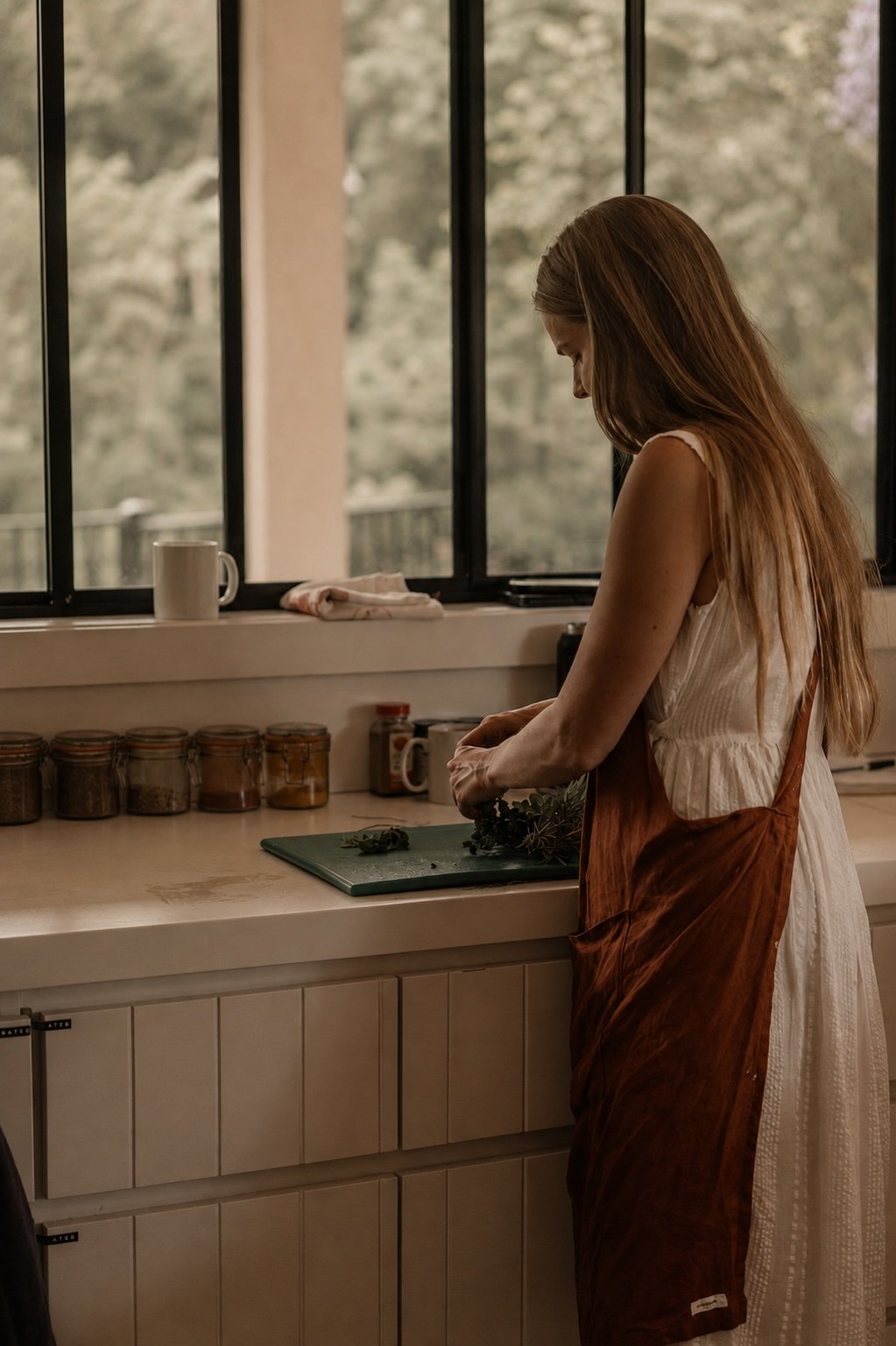 A woman with long reddish hair, wearing a sleeveless white dress and an orange apron, standing at a kitchen counter near a window. She appears to be preparing herbs or greens on a cutting board. The background shows a scenic view of trees outside, and the counter has jars and a coffee mug.