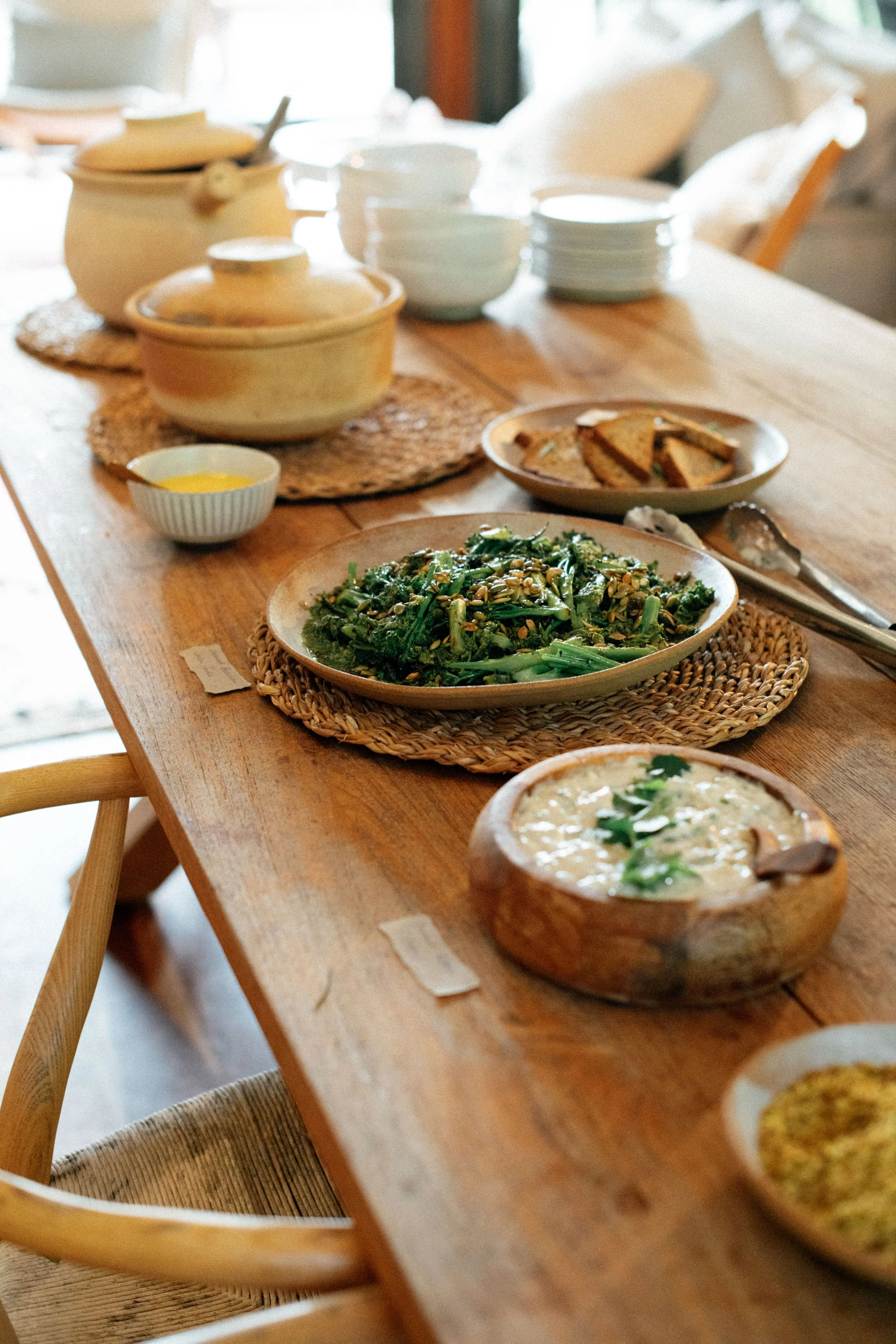 A rustic dining table set with various dishes including a green vegetable salad, a bowl of creamy soup, and plates of bread, with stacks of white bowls and a yellow sauce on the side.