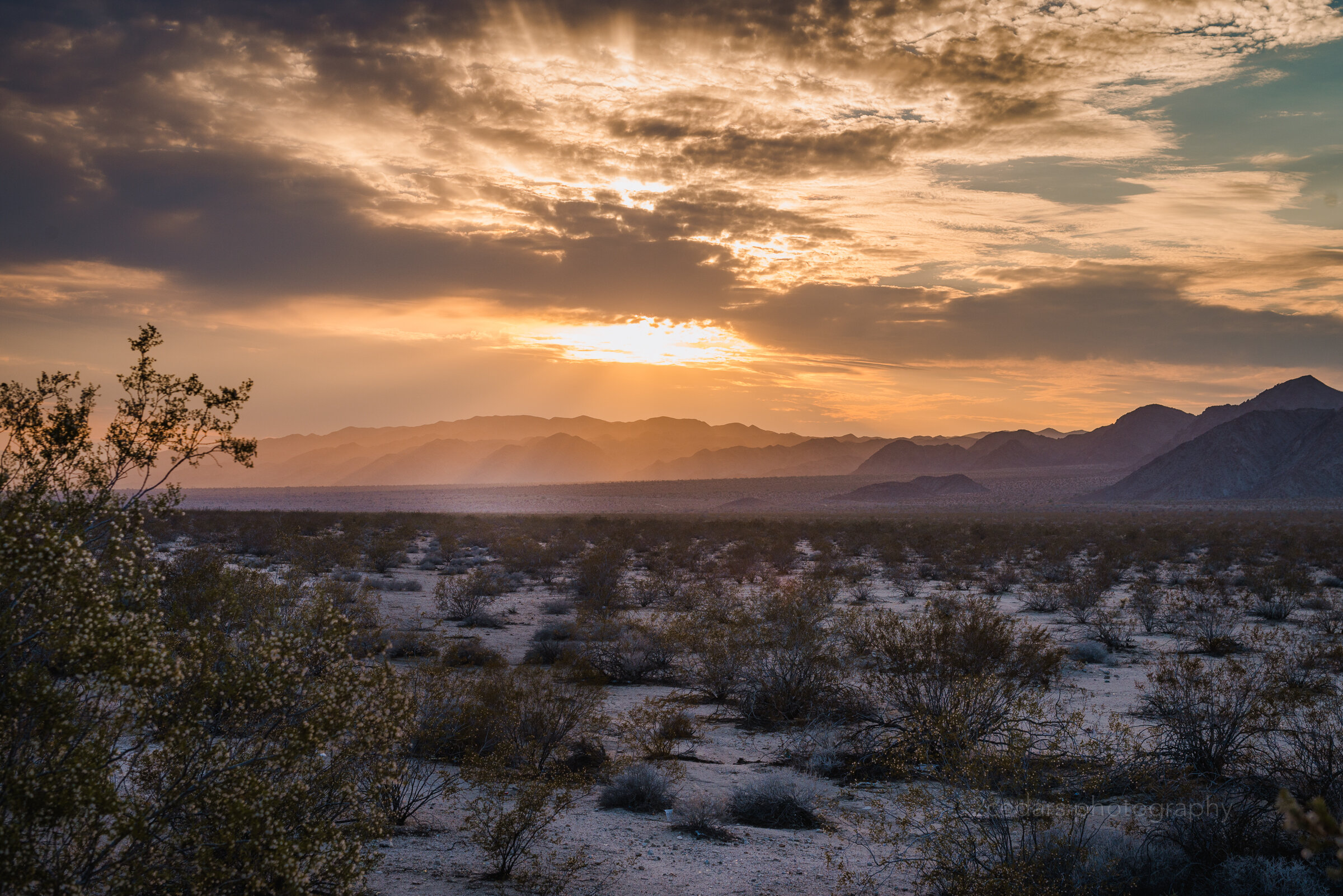  A sunset streaming through the clouds over a white sand and creosote bush desert just behind the hills in the background.  