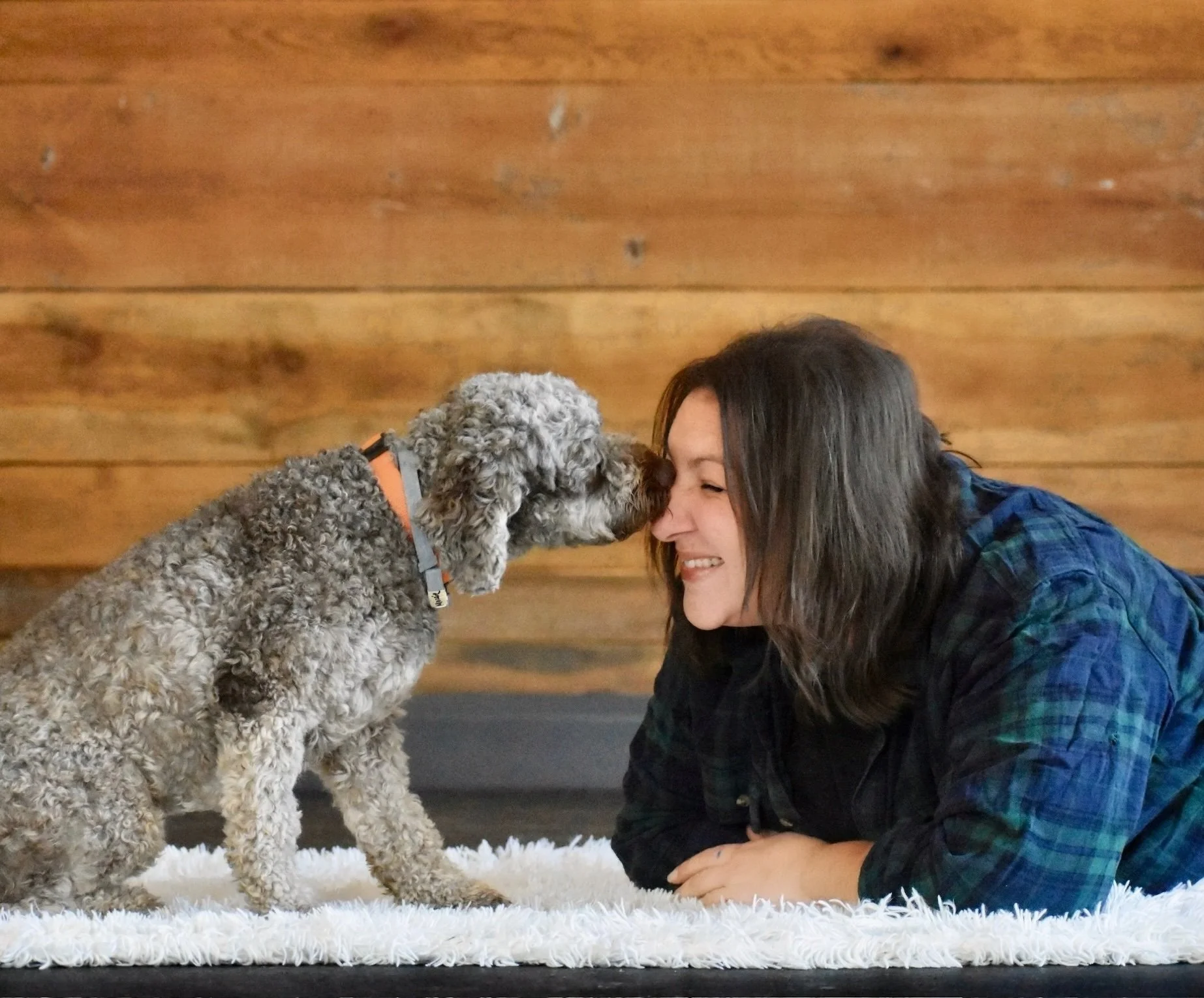 Melissa Ragaini, Team Lead at Far Fetched Acres, lying on the floor indoors as a dog affectionately nuzzles her face.