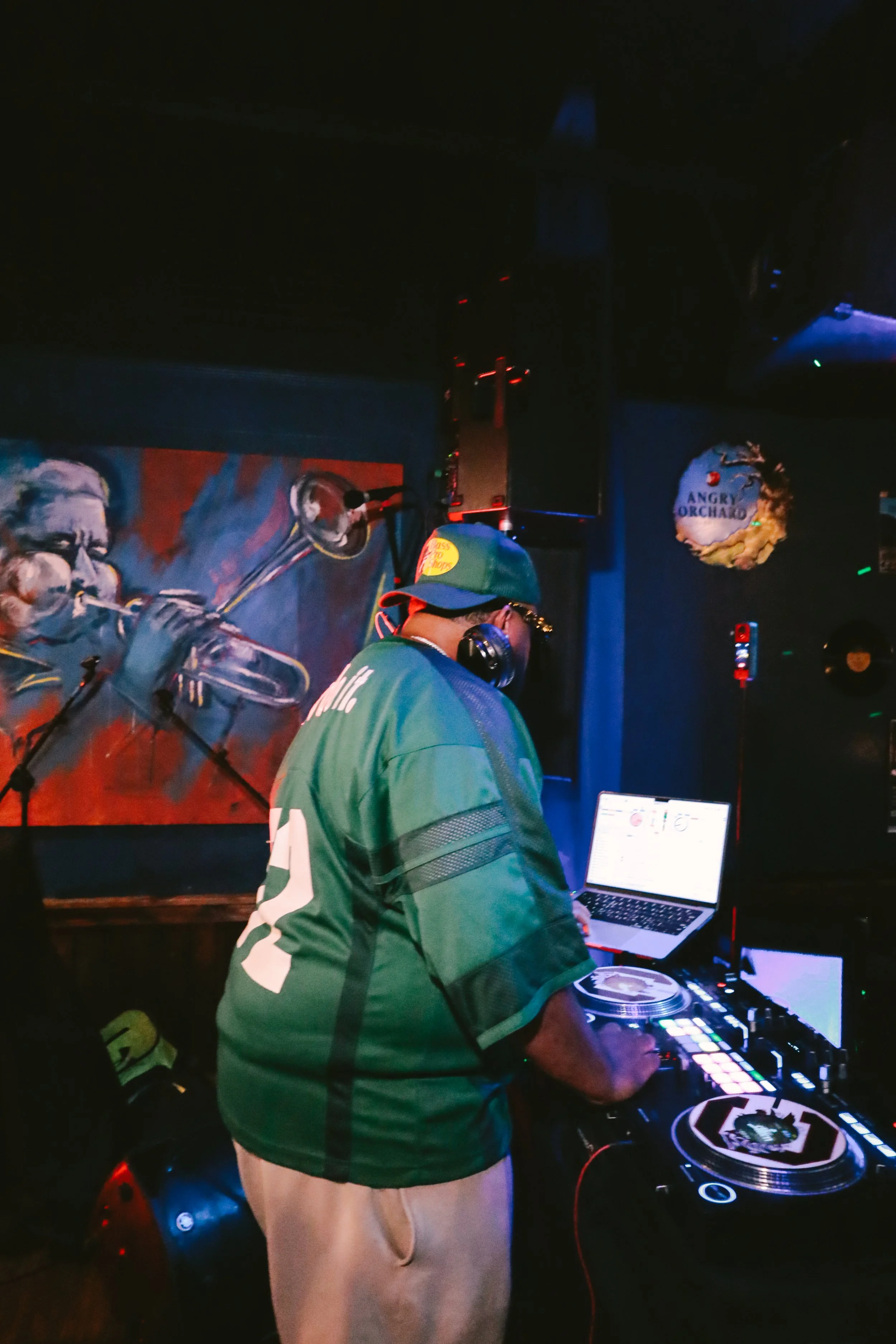 A DJ wearing a green jersey and beige pants operates a DJ mixer with headphones in a dimly lit room. A skateboard is placed on the floor nearby. There is a colorful mural of a man playing a trumpet on the wall behind him, and part of a sign that read