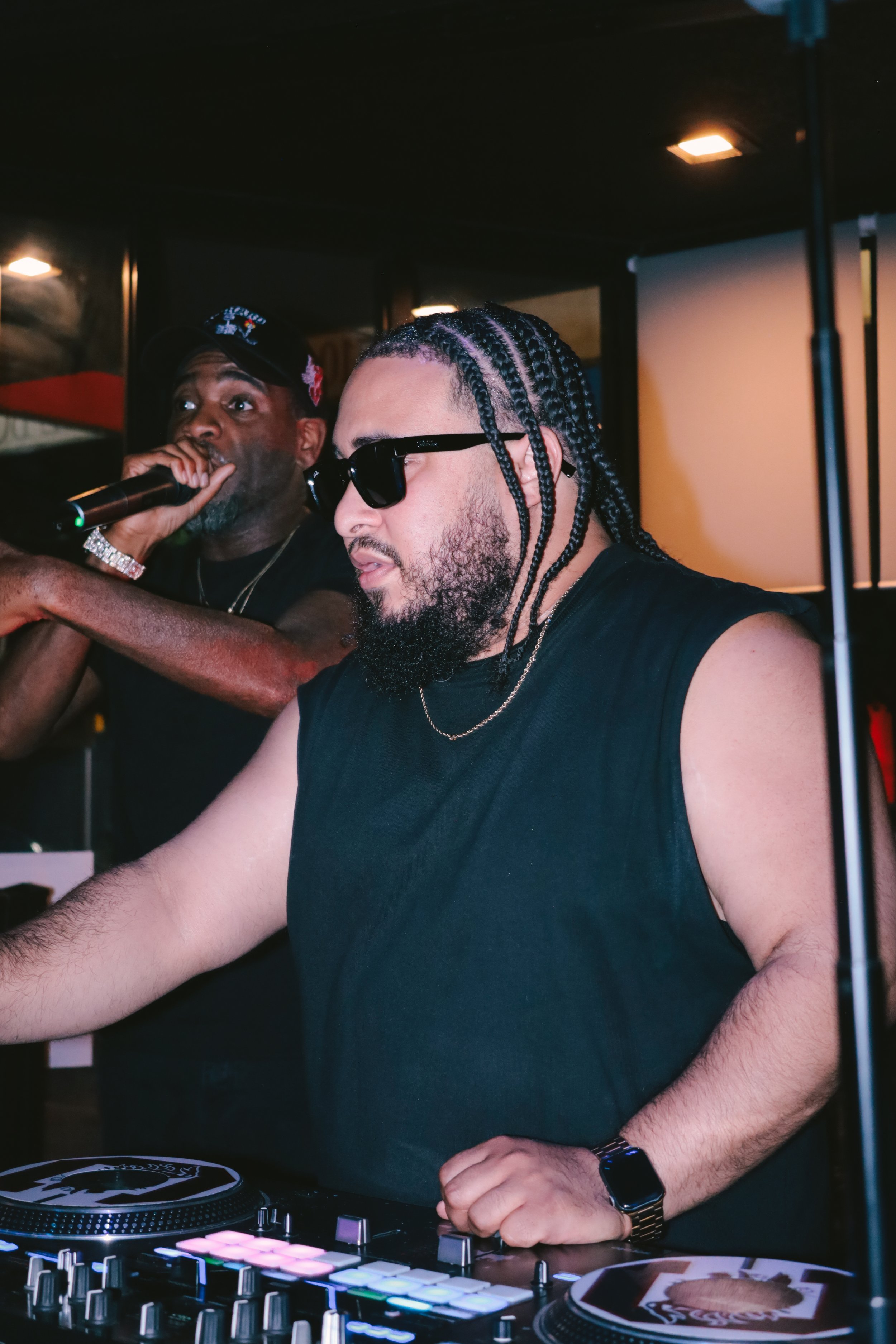 A DJ with braids, sunglasses, a beard, wearing a black sleeveless shirt, using DJ equipment with colorful pads in a club or music venue.