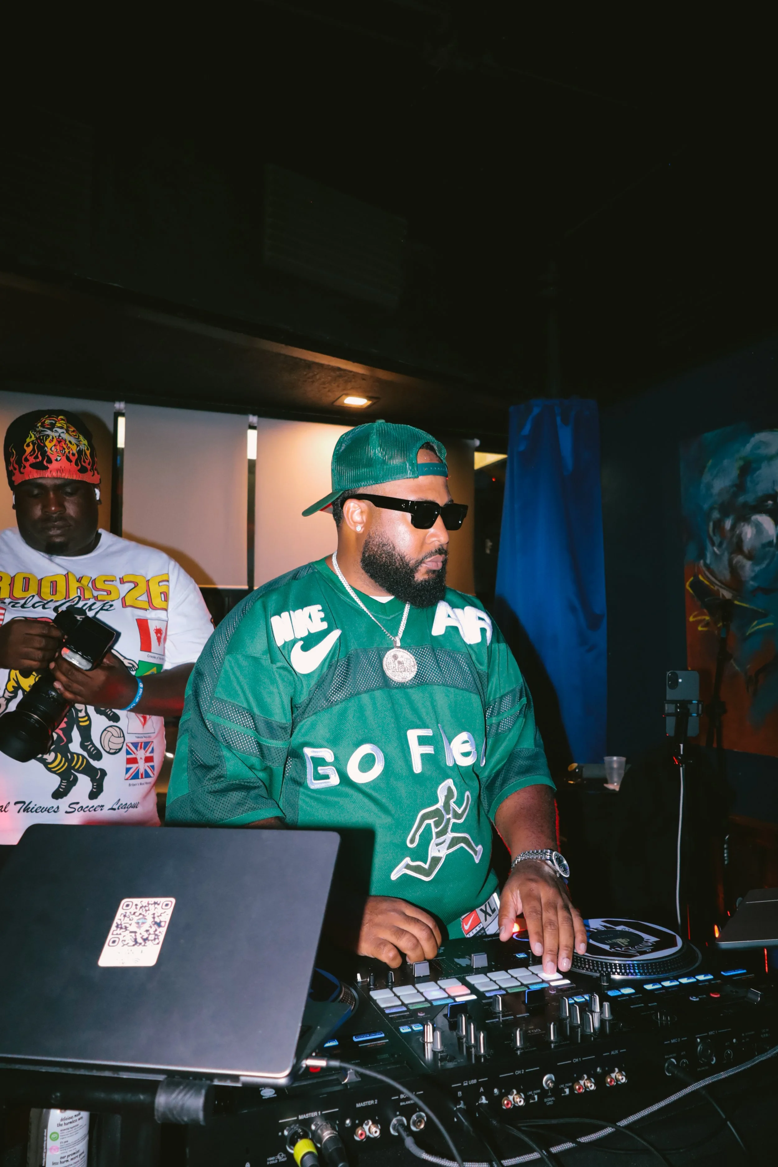 DJ iAM is mixing music at a nightclub, wearing a green football jersey, a green cap, and sunglasses, with a DJ mixer in front of him. There are other people and artwork in the background.