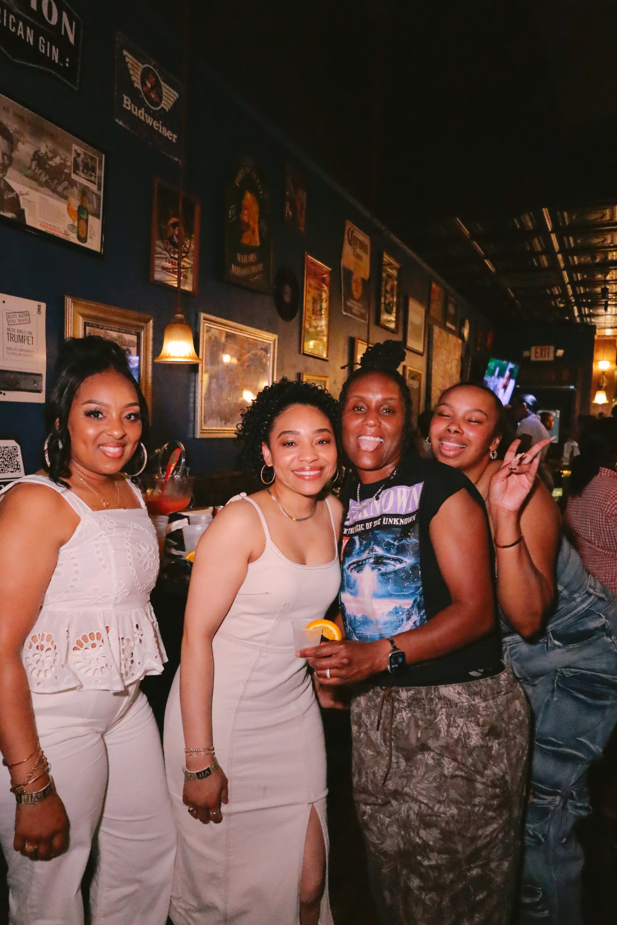 Four women standing together in a bar, smiling for the camera. The wall behind them is decorated with framed pictures, posters, and vintage signs, including poster advertisements and a Budweiser logo. One woman is making a peace sign, another holds a