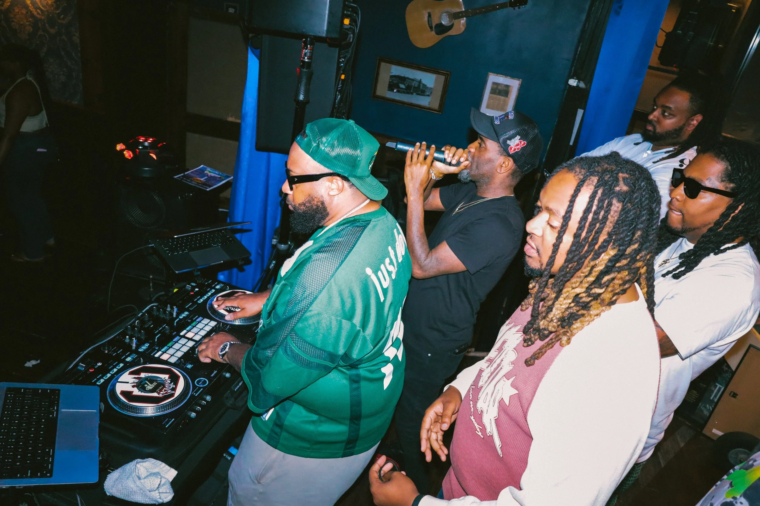 A group of men at a DJ setup with music equipment, some with dreadlocks, one wearing sunglasses, and another in a baseball cap using a microphone. They appear to be in a dimly lit indoor space with framed pictures on the blue wall behind them.