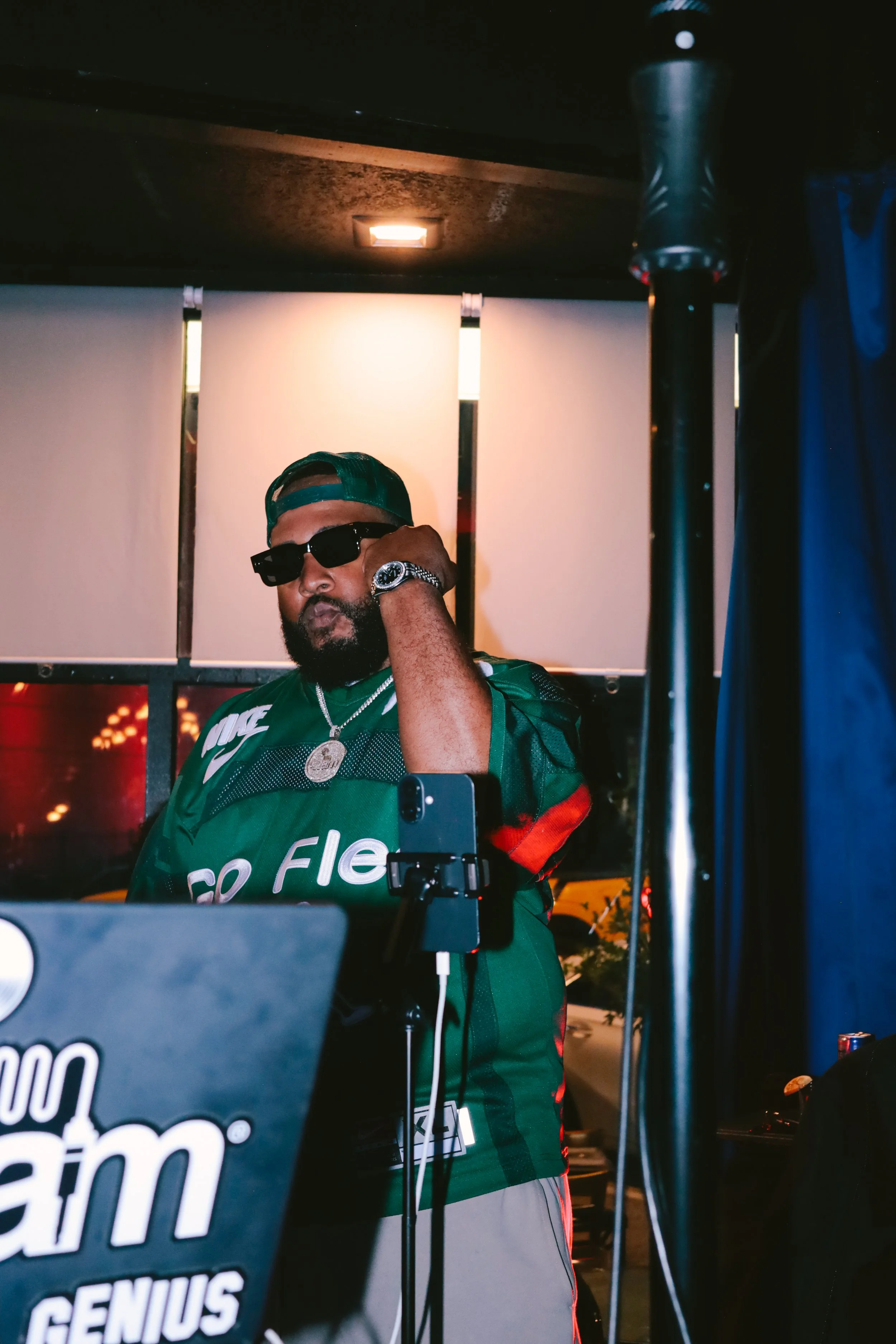 DJ iAM wearing a green jersey, sunglasses, and a cap, standing behind DJ equipment in a dimly lit indoor setting.