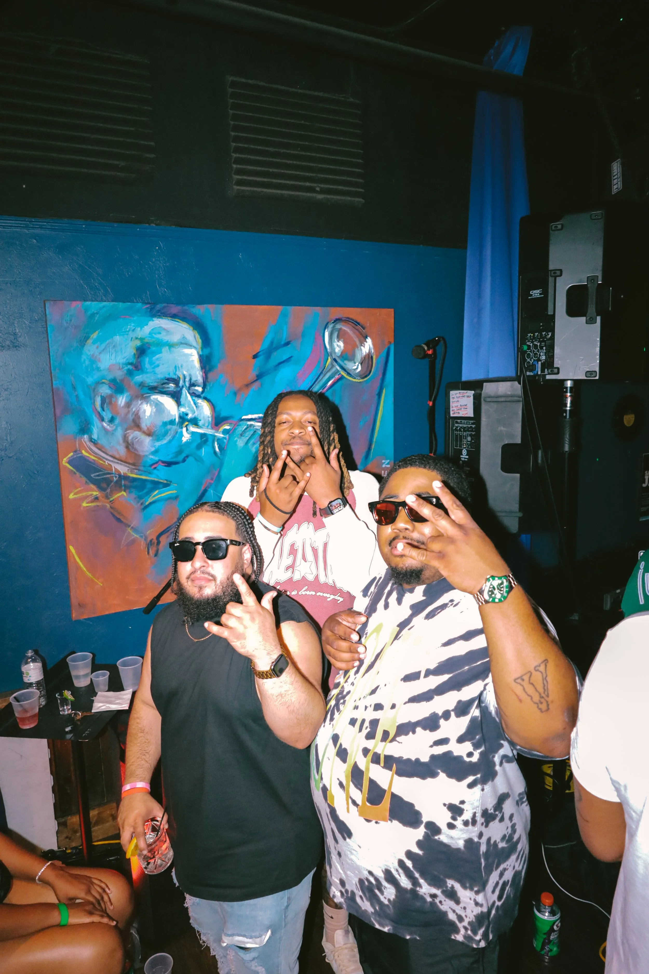 Group of people at a party or social gathering, posing for a photo near a colorful painting of a man playing a trumpet, with DJ equipment and drinks on a table in the background.