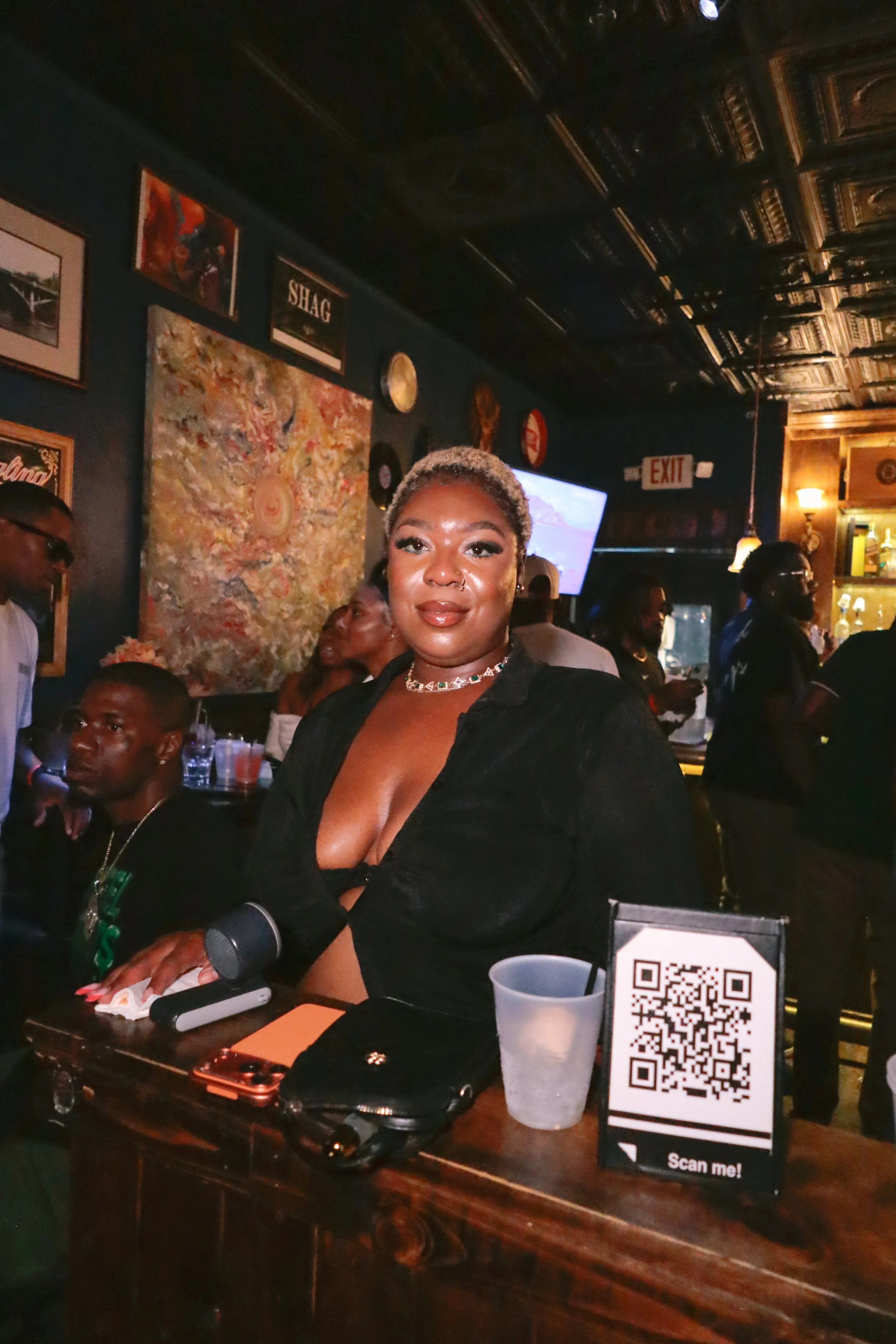 A woman with short blonde hair and a nose piercing is sitting at a table in a dimly lit bar or restaurant, smiling at the camera. The table has a QR code stand, a drink cup, a smartphone, and a microphone. There are people in the background, wall art