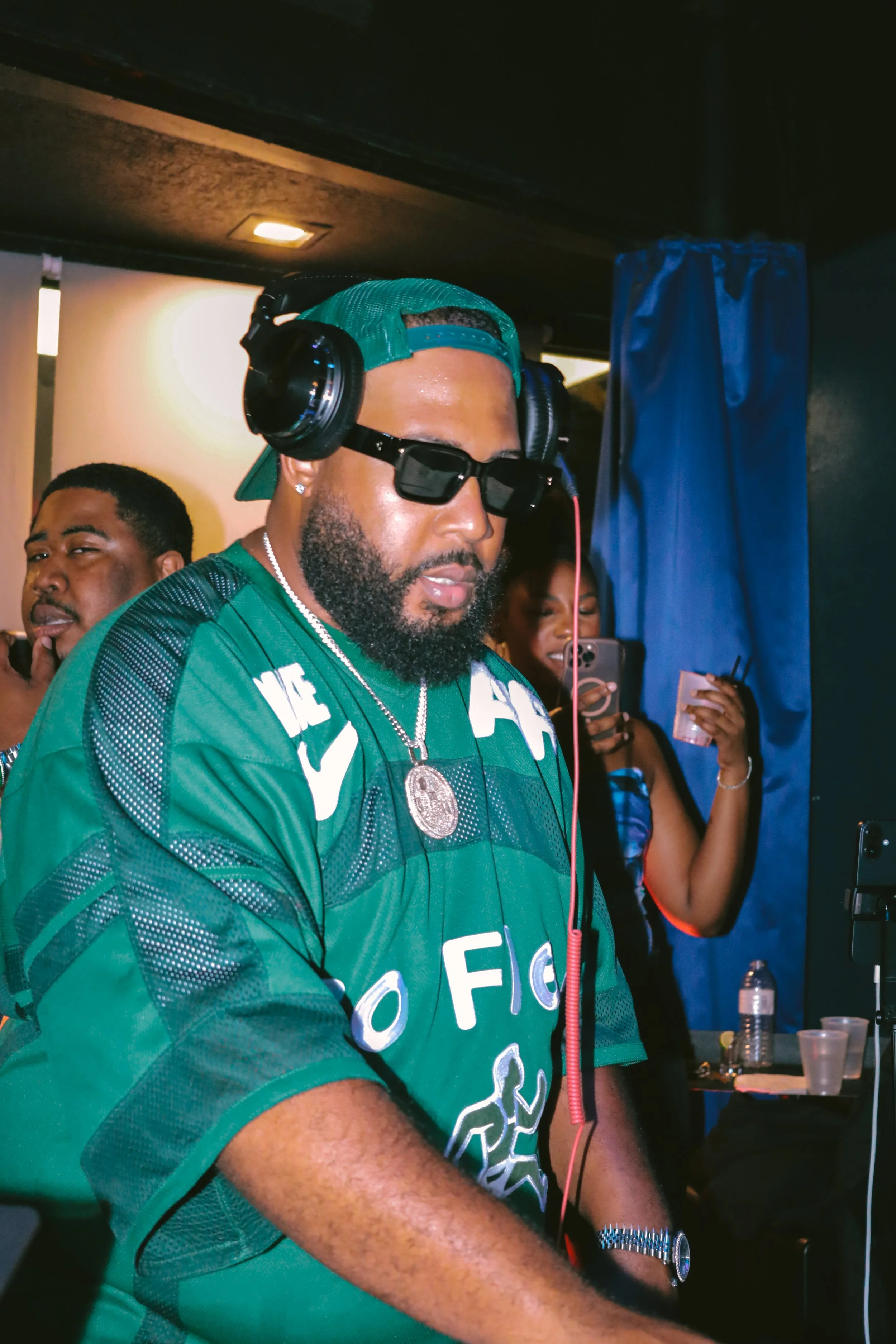 A man with a beard wearing sunglasses, a green cap backward, and a green sports jersey, standing indoors with people in the background. He is listening to music through over-ear headphones, with a serious expression.