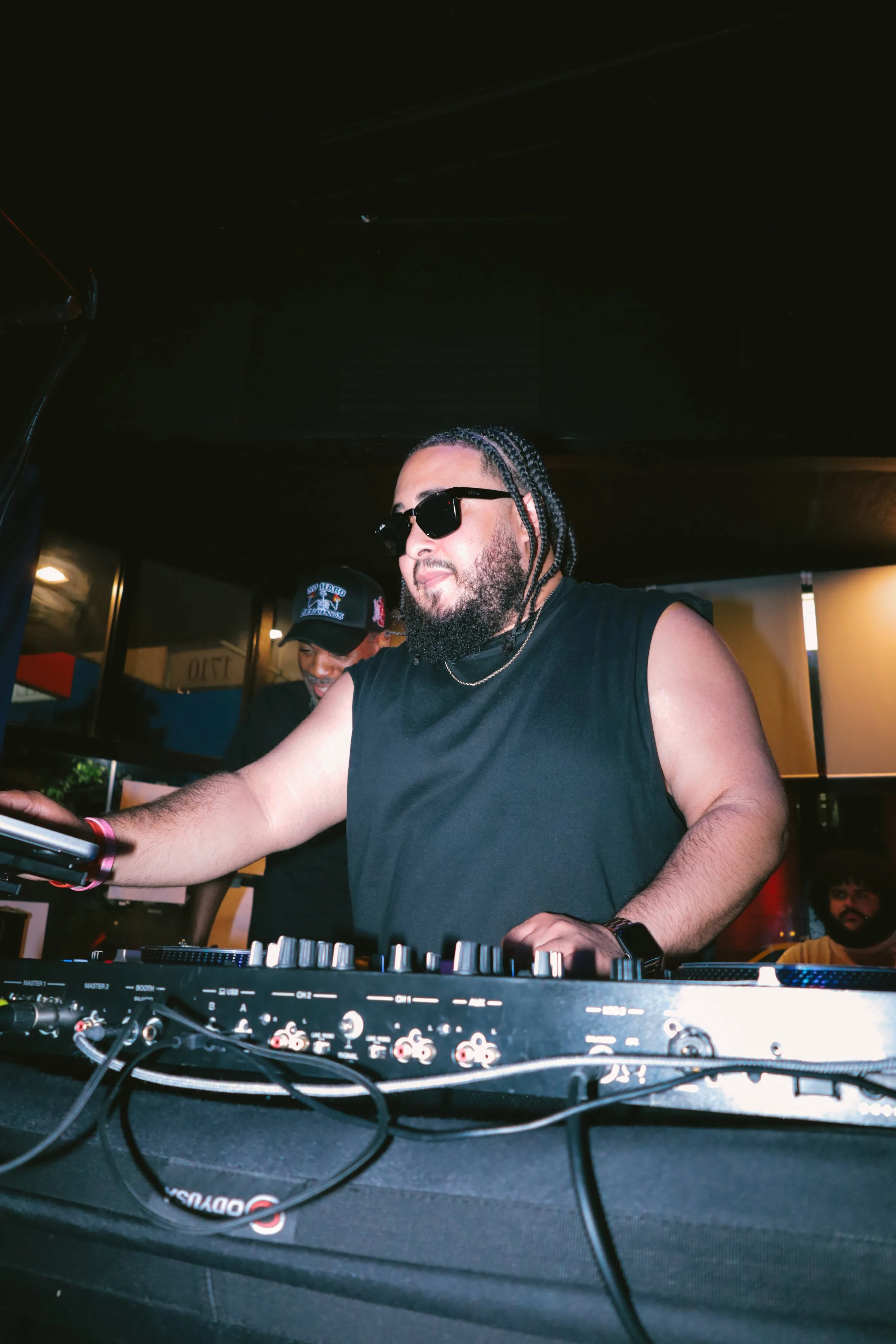 A man with braided hair, sunglasses, and a beard, wearing a black sleeveless shirt, DJing at a music event with DJ equipment, and a person in a cap in the background.