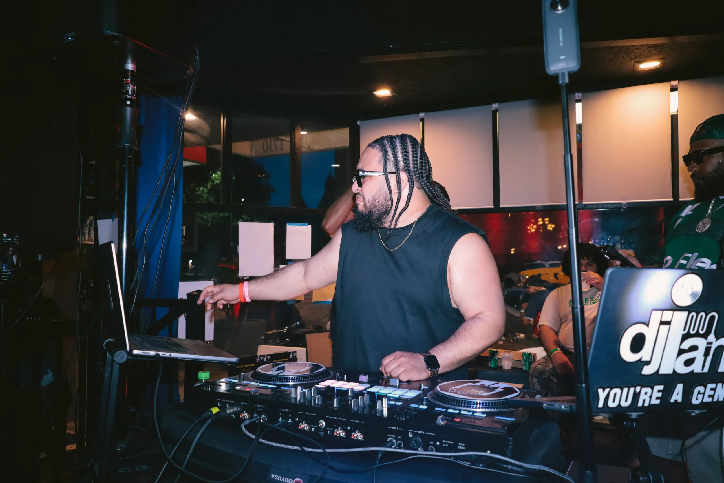 A DJ with braided hair and sunglasses playing music at a nightclub with people in the background.