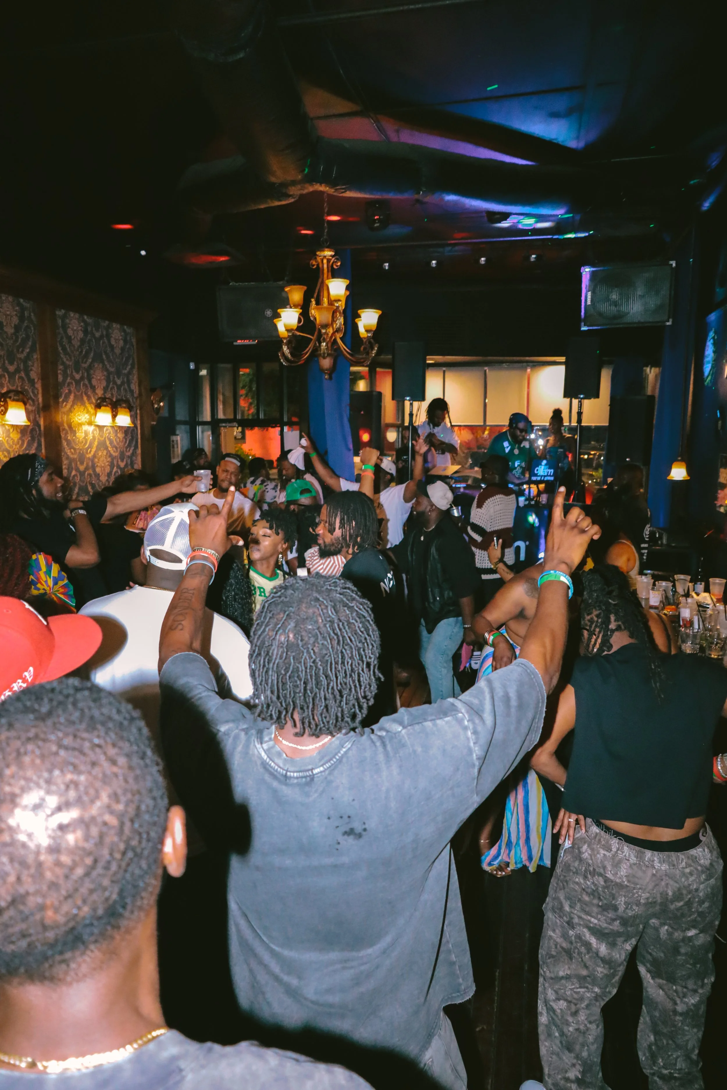 People dancing and socializing in a nightclub, with DJ playing music, dim lighting, and colorful stage lights.