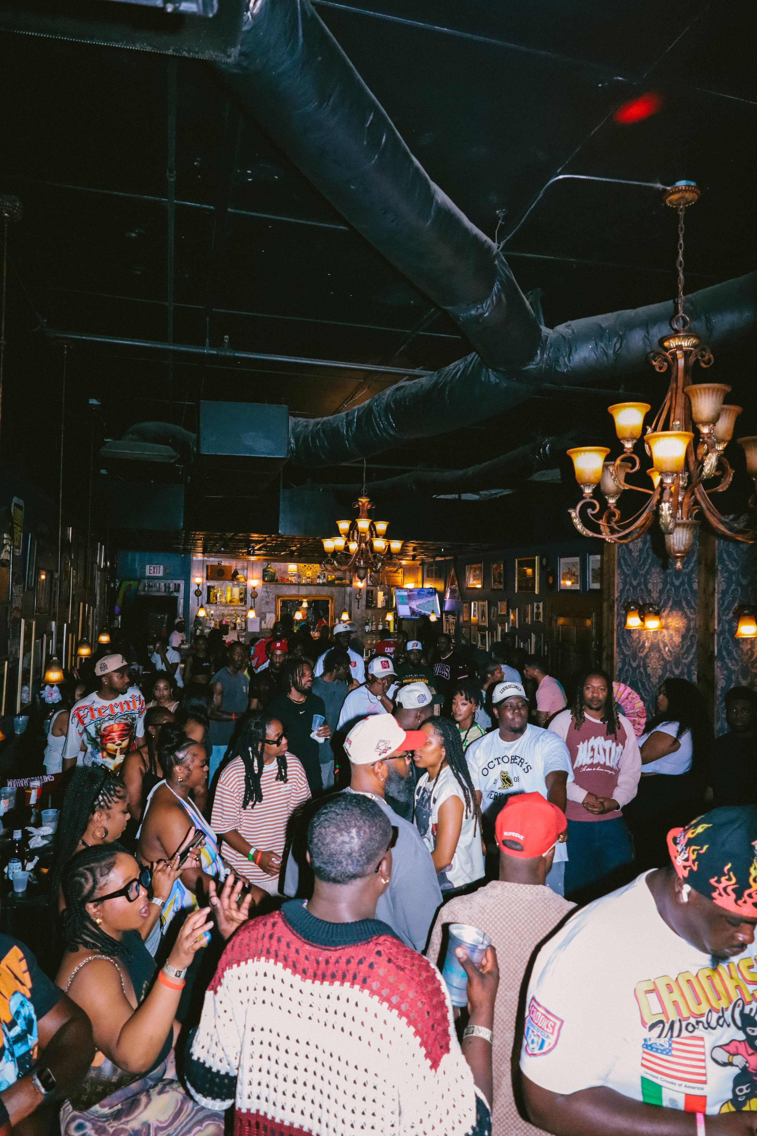 Crowd of people gathered indoors at a bar or club, with black ceiling pipes and ornate chandeliers hanging from the ceiling.