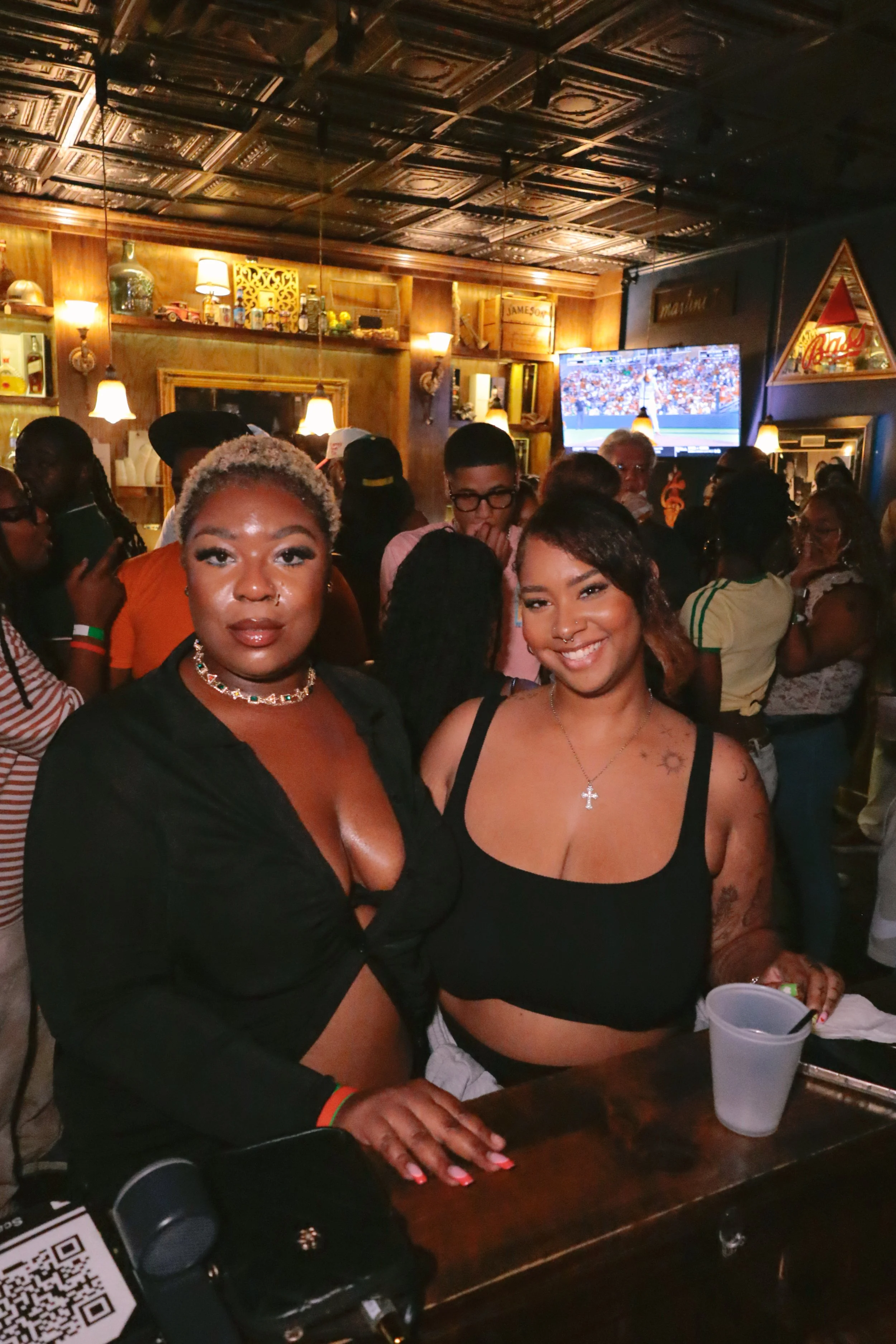 Two women with dark skin and tattoos on their arms standing at a bar in a crowded pub, smiling and looking at the camera, with a large TV screen showing a sports game in the background.
