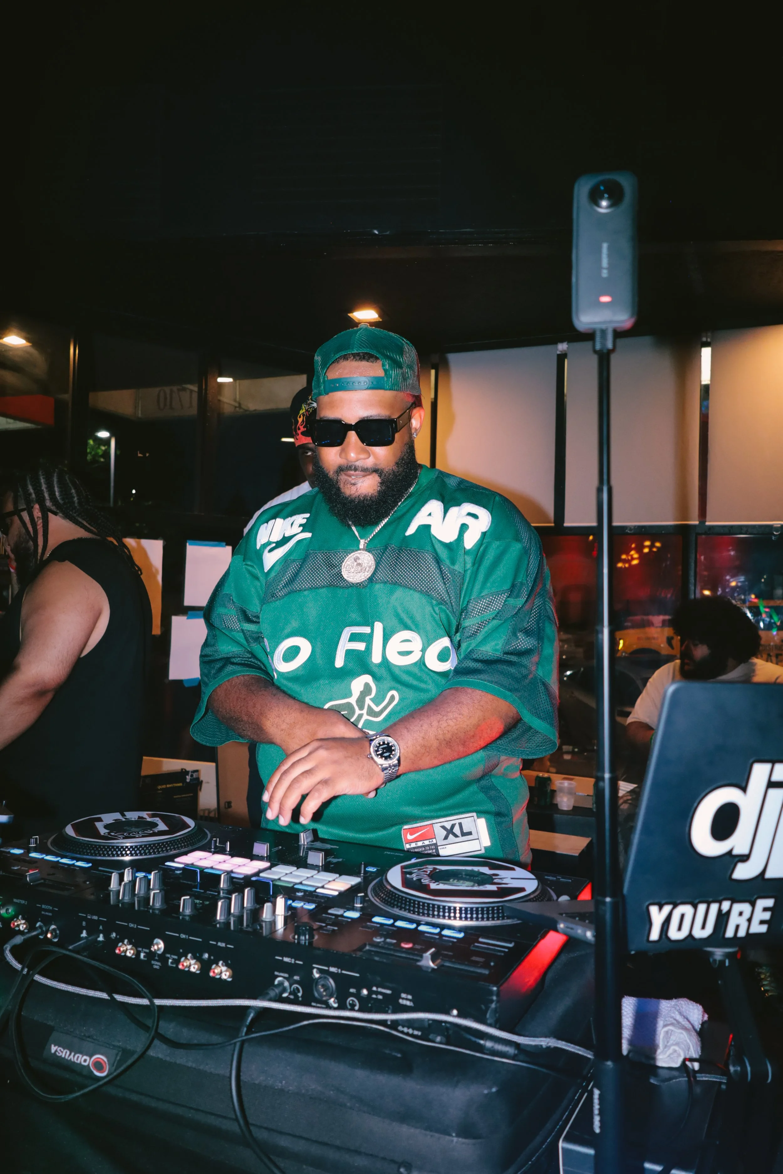 A DJ wearing sunglasses, a green sports jersey, a green cap, and a large necklace, standing behind a DJ mixer at a party or event. The surroundings include people and equipment.