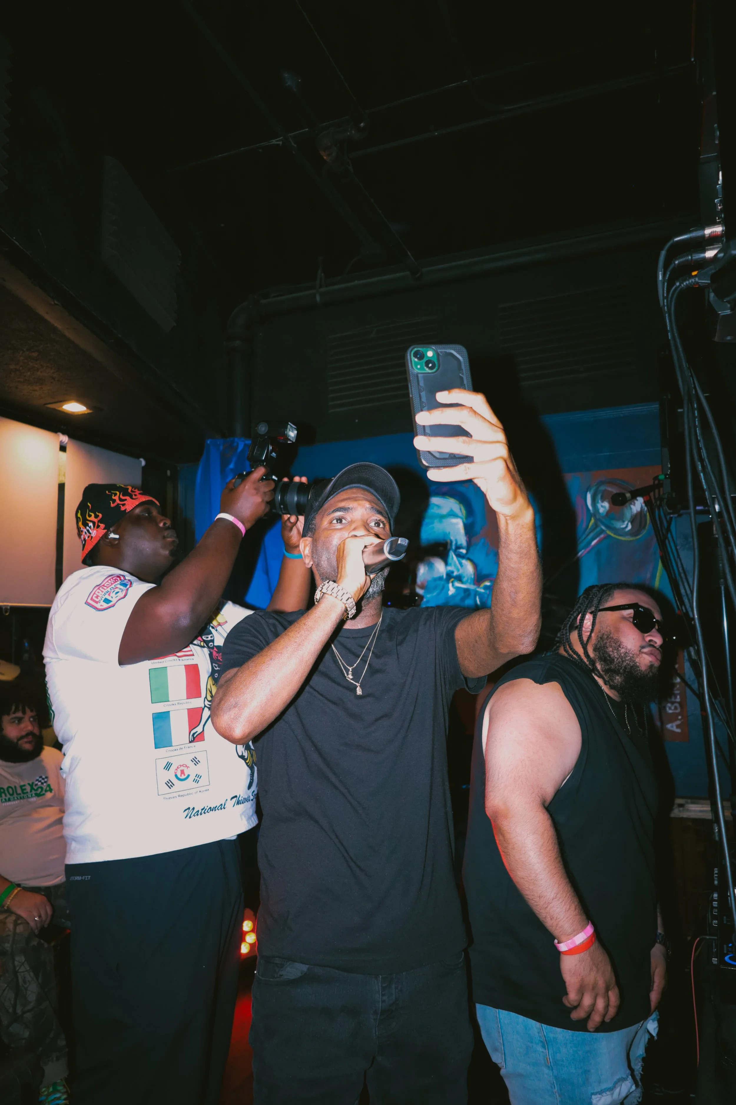 People at a music event with one person taking a selfie, others in black and white tank tops, one wearing a cap, and colorful lighting in the background.
