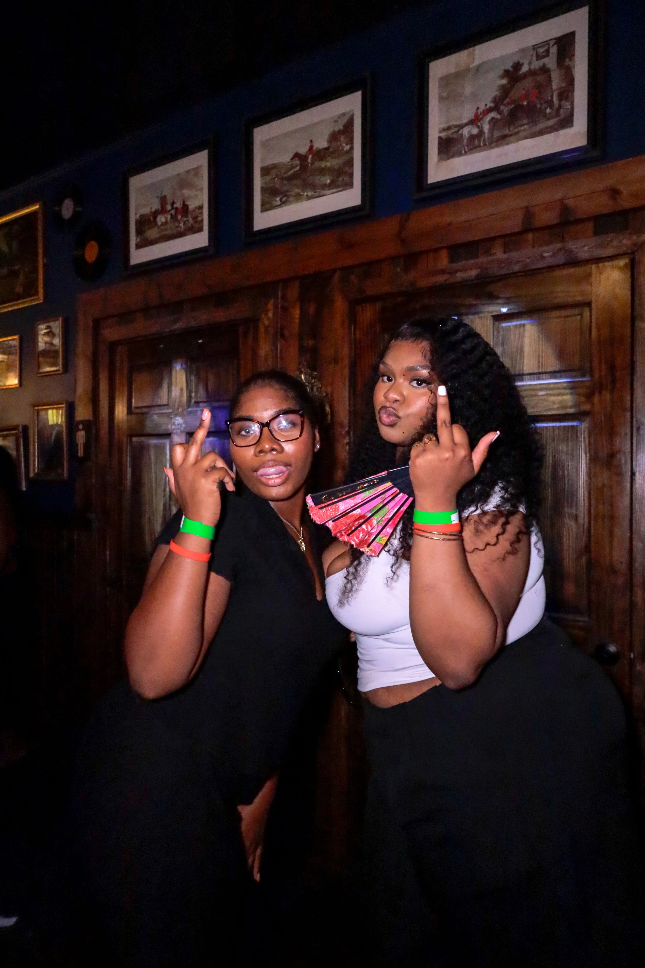 Two women are posing with serious expressions, holding up their middle fingers. They are in a dimly lit room with wooden walls and framed art or photos hanging above them.