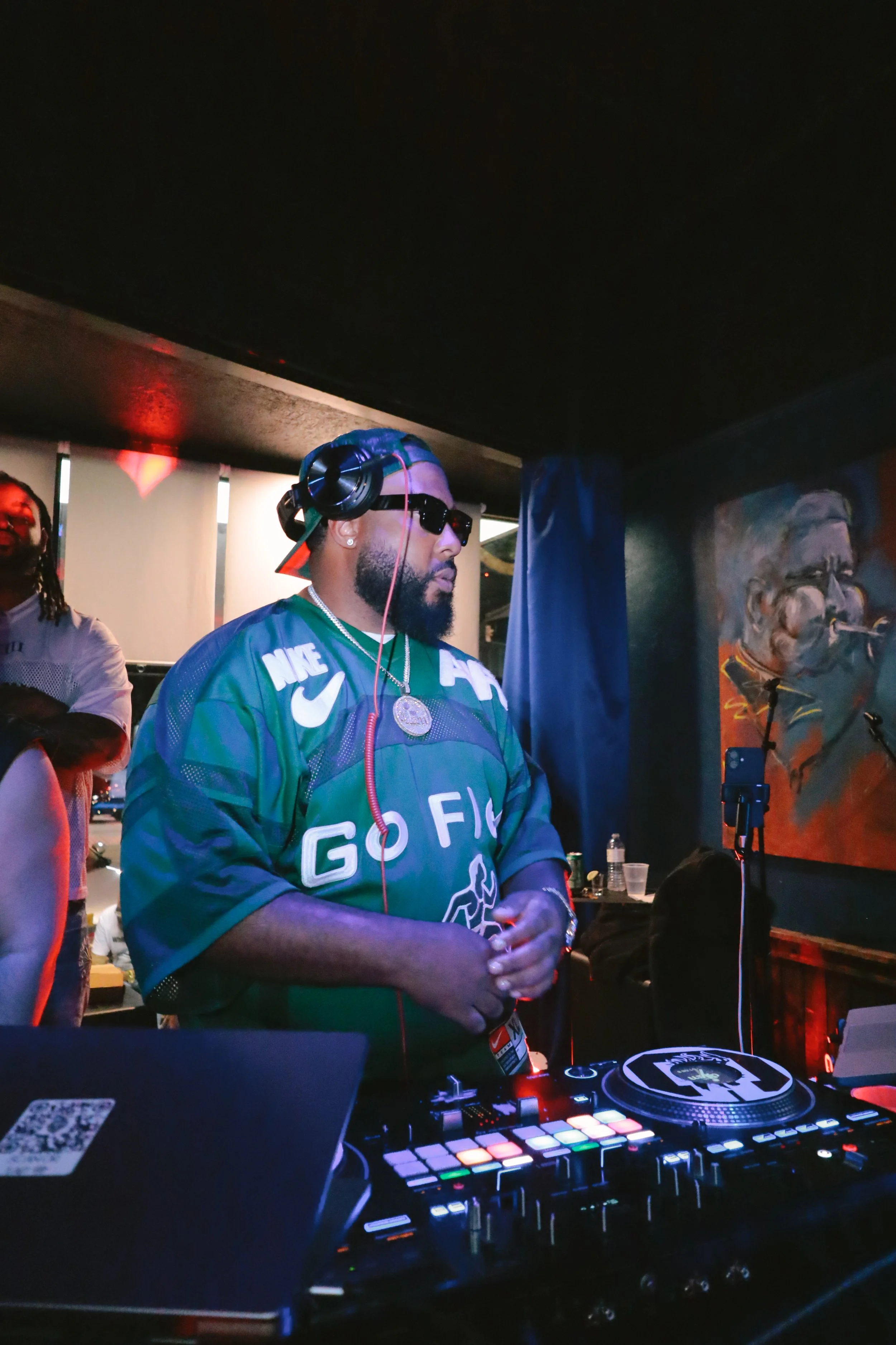 A DJ wearing sunglasses, a cap backward, and a green Nike jersey is performing at a nightclub or music event. He is using a DJ controller with colorful pads, with a painting of a man singing on a microphone in the background.
