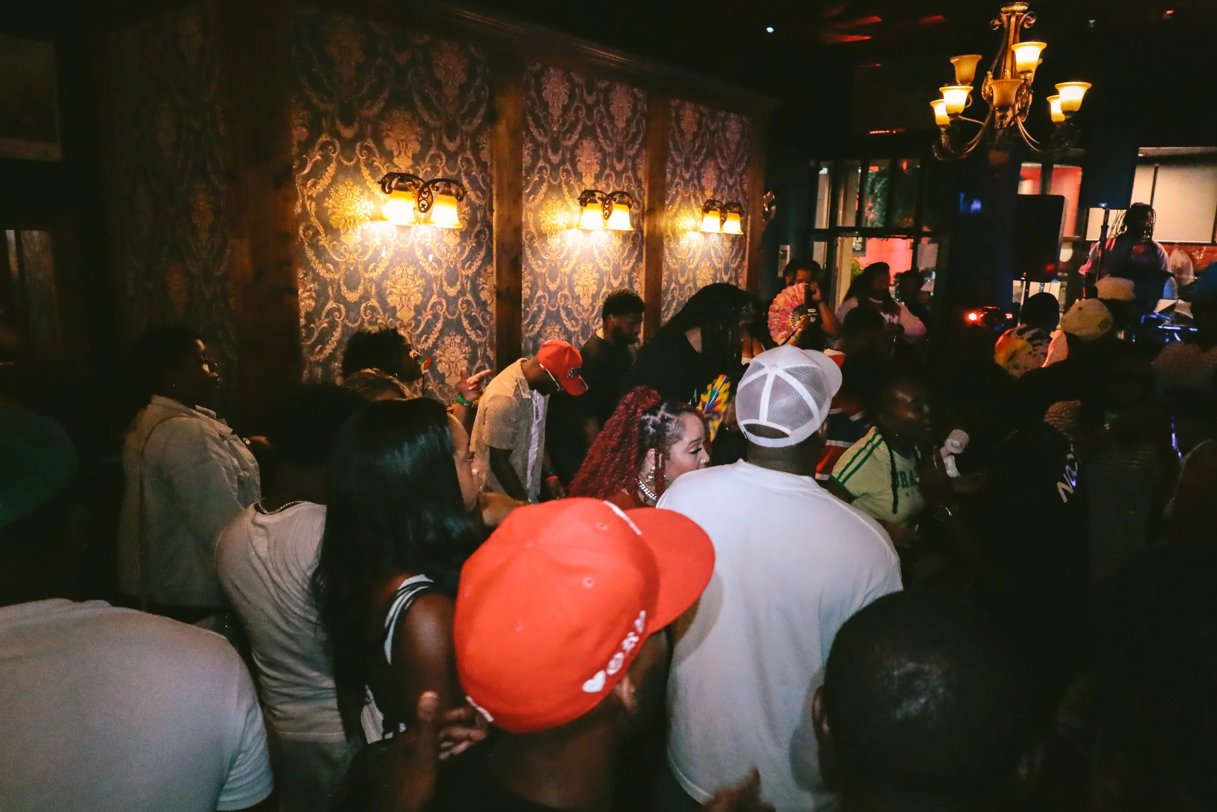 Crowd of people gathered in a dimly lit room with ornate wallpaper and vintage-style wall lamps, possibly at a concert or event.