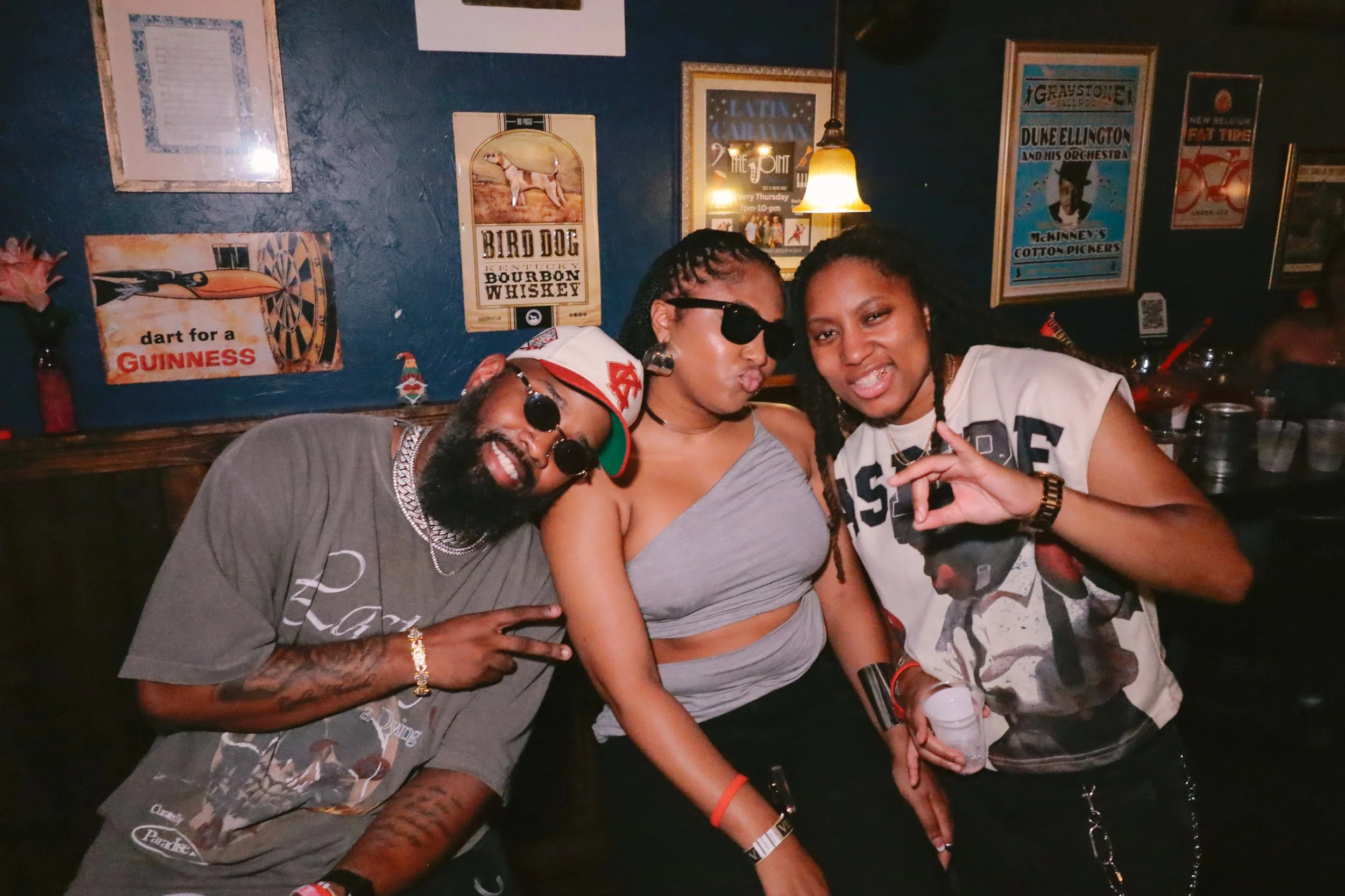 Three friends posing together at a bar with vintage posters on the wall behind them. They are smiling and making gestures towards the camera. One is wearing sunglasses, and another is holding a drink.