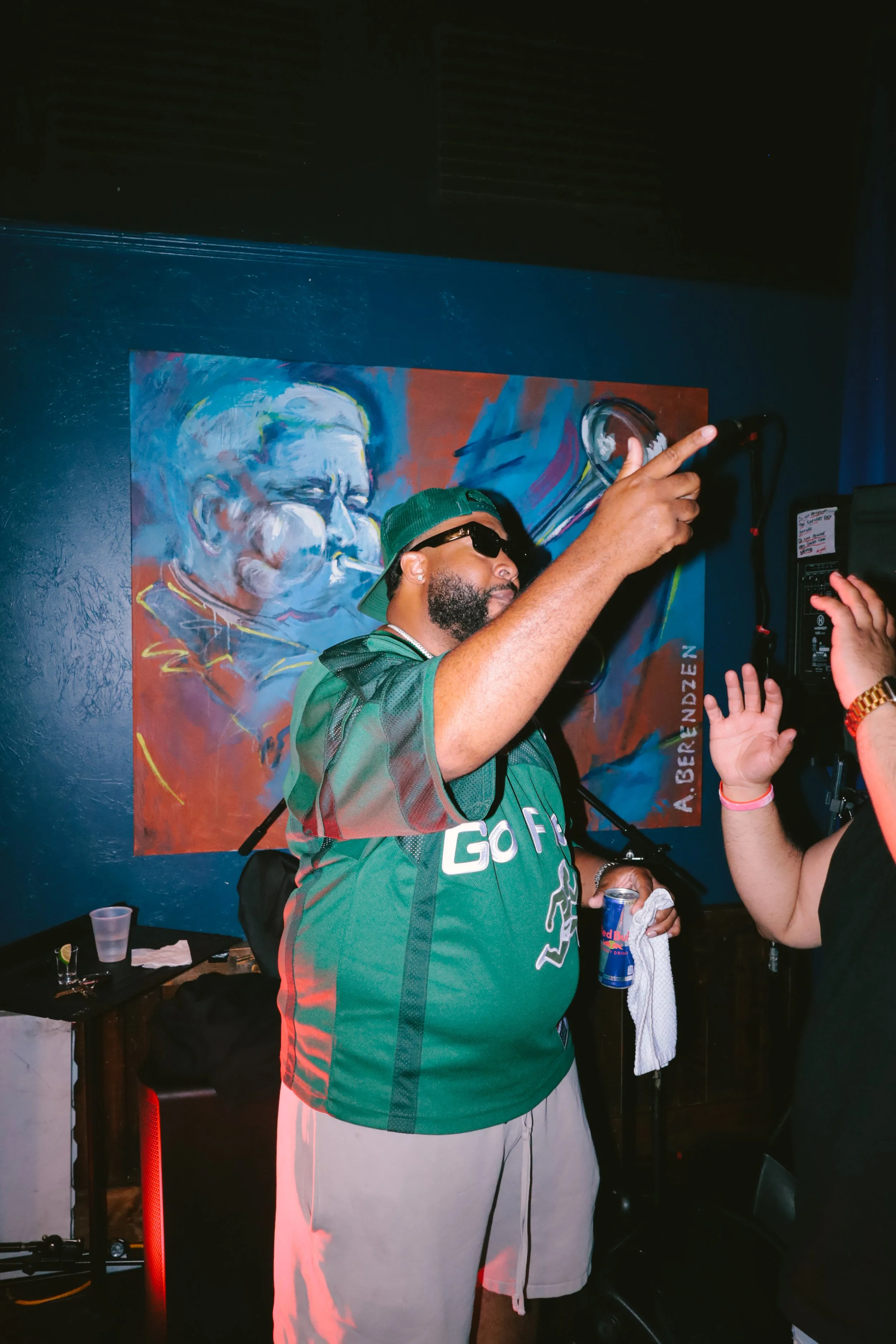 A man wearing sunglasses, a green cap, and a green sports jersey with the word 'GO' on it, holding a Red Bull can and a towel, standing in front of a large painting of an elderly man smoking.