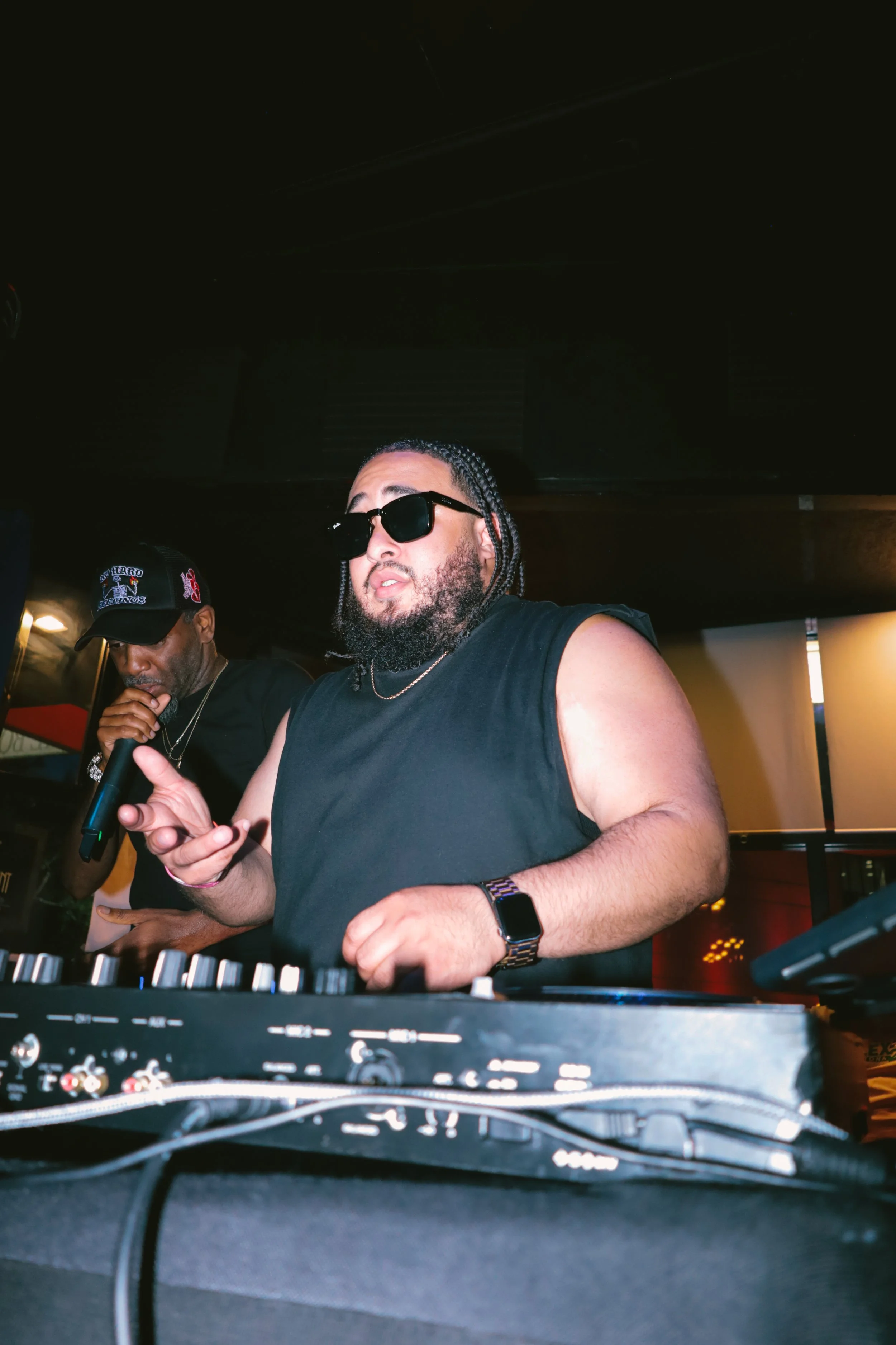 A DJ wearing sunglasses and a sleeveless shirt performing with two men in the background, one with a cap, in a dimly lit indoor venue.