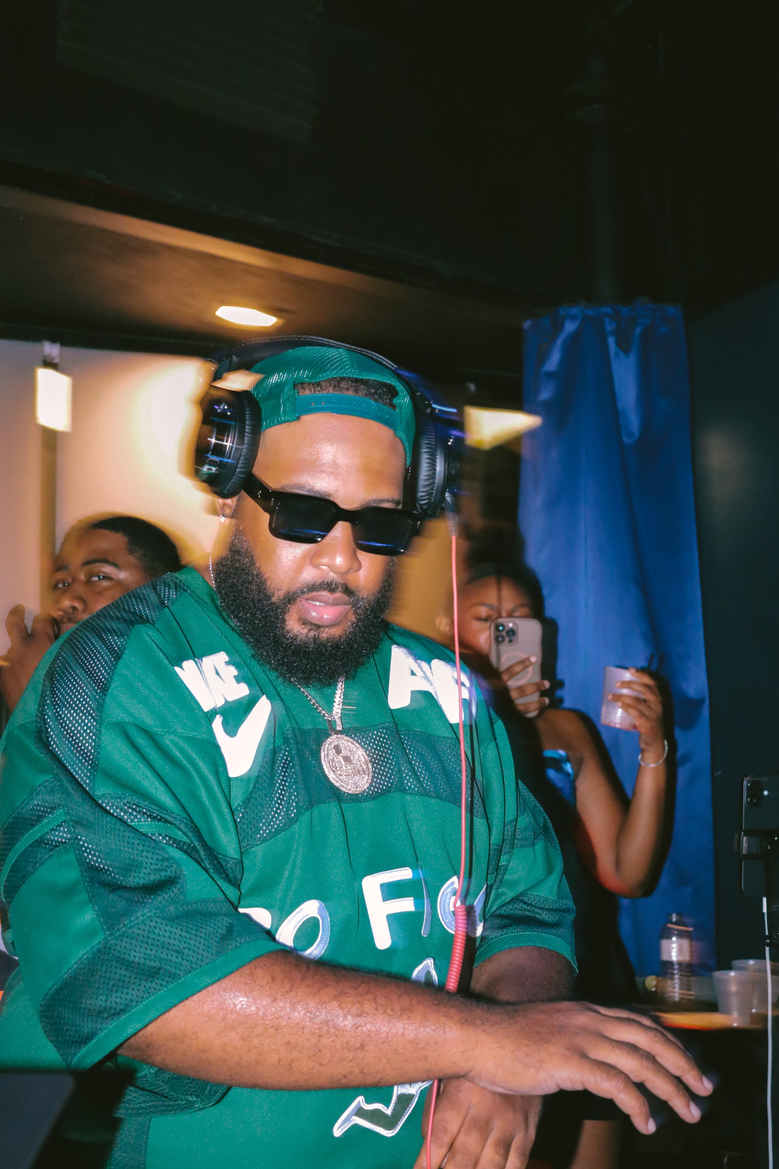 A man wearing a teal snapback, sunglasses, and a green sports jersey with white letters and a large silver pendant, appears to be DJing or performing music with headphones and a red cable, with people in the background taking photos.