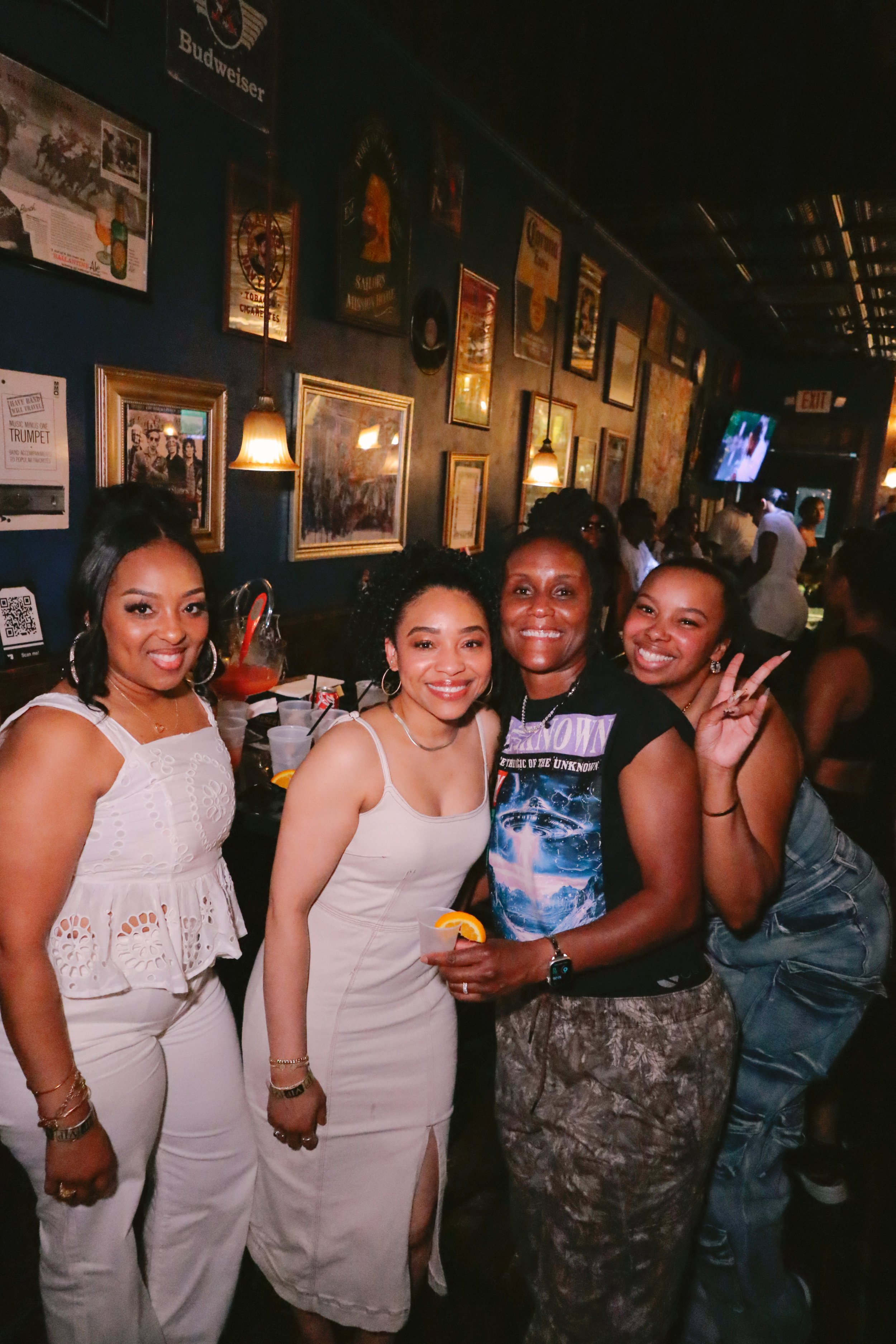 Four women standing together at a bar or restaurant, smiling, with framed pictures and posters on the dark wall behind them, and a television screen in the background showing a sports event.