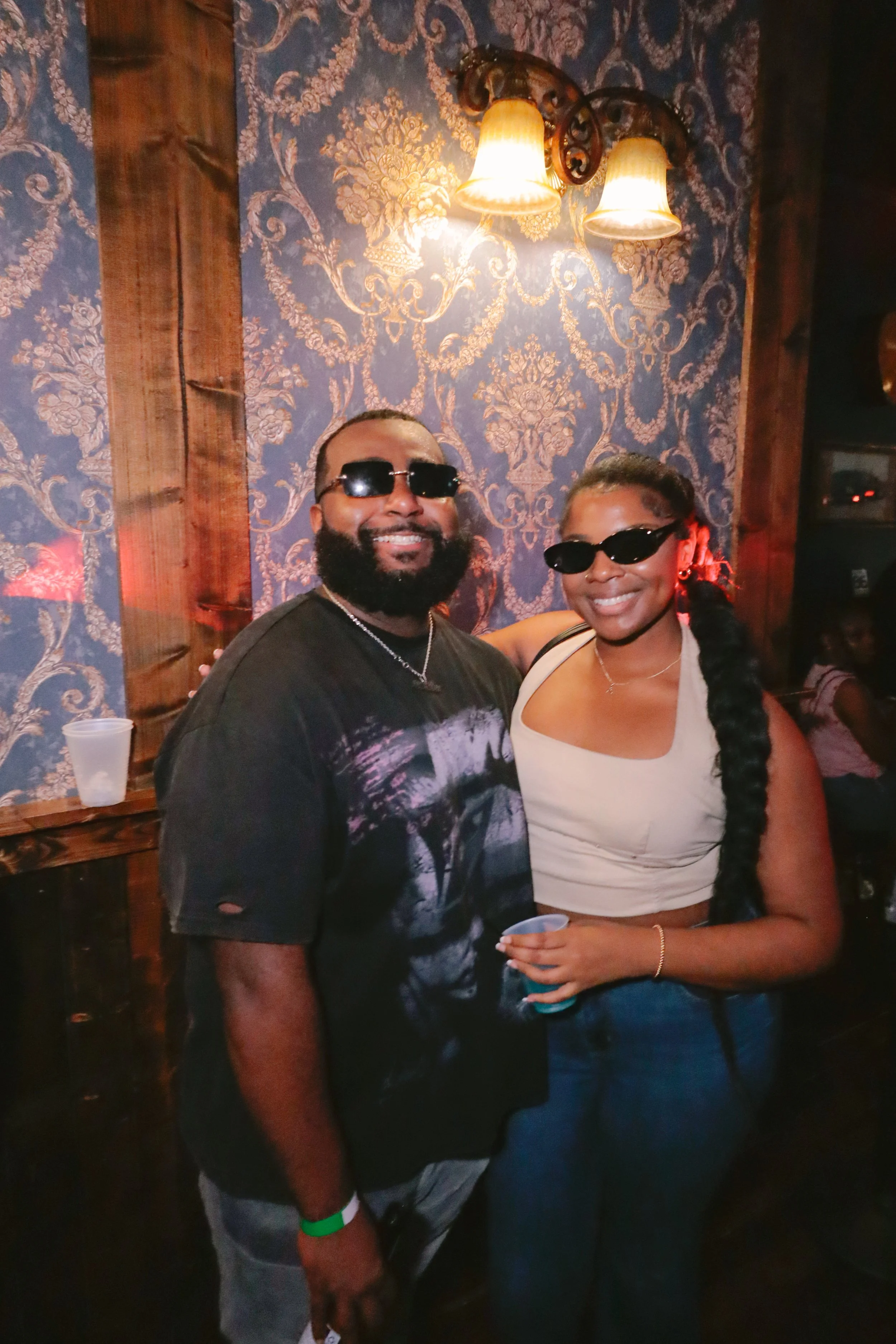 A man and woman wearing sunglasses stand close together, smiling at the camera in a dimly lit room with ornate wallpaper and a wall-mounted light fixture. The man has a beard, wears a dark graphic T-shirt, and a green wristband. The woman has braided