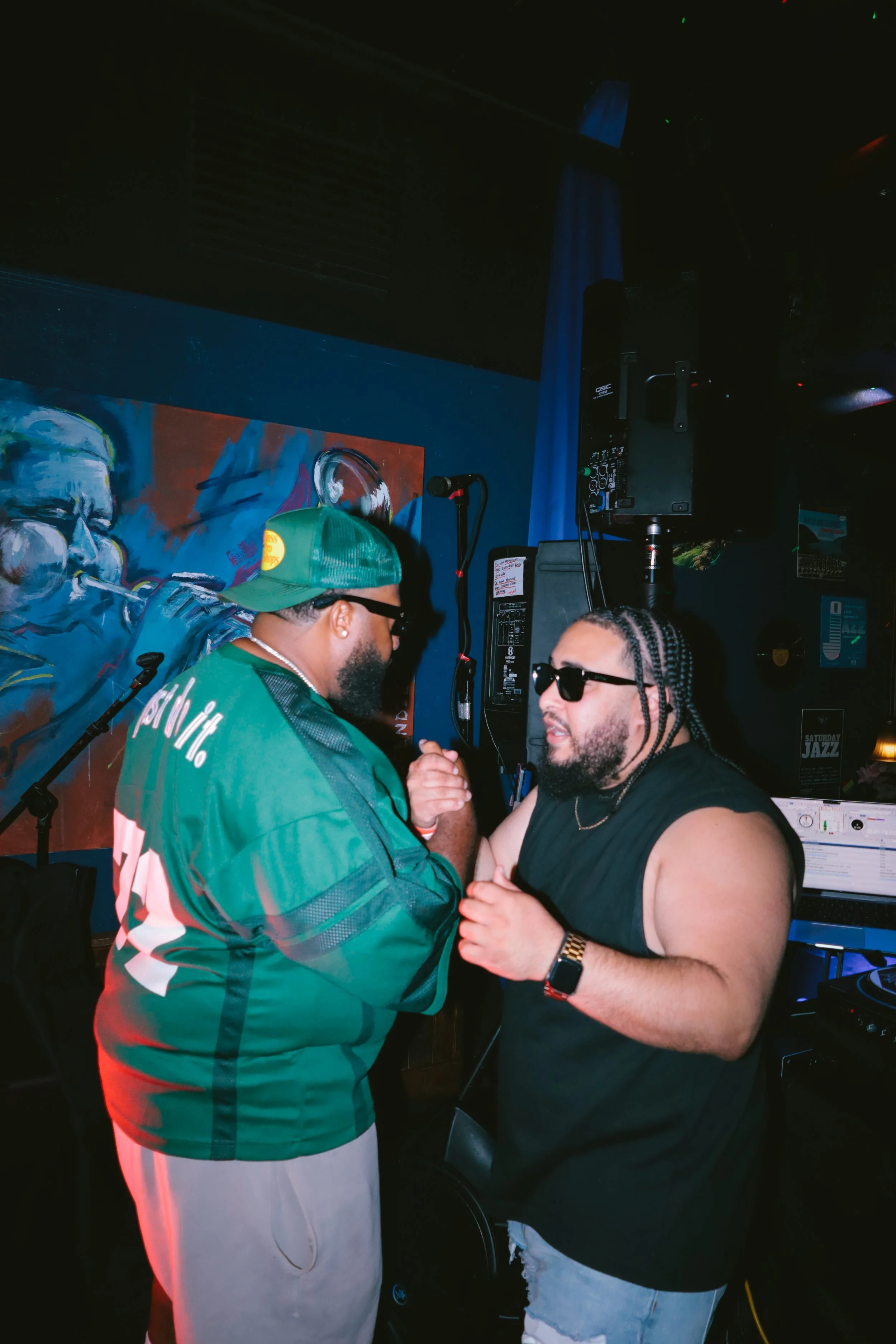 Two men are dancing closely at a nightclub, with one in a green jacket, white pants, and a green cap, and the other in a black tank top and sunglasses. The background features a colorful painting and DJ equipment.
