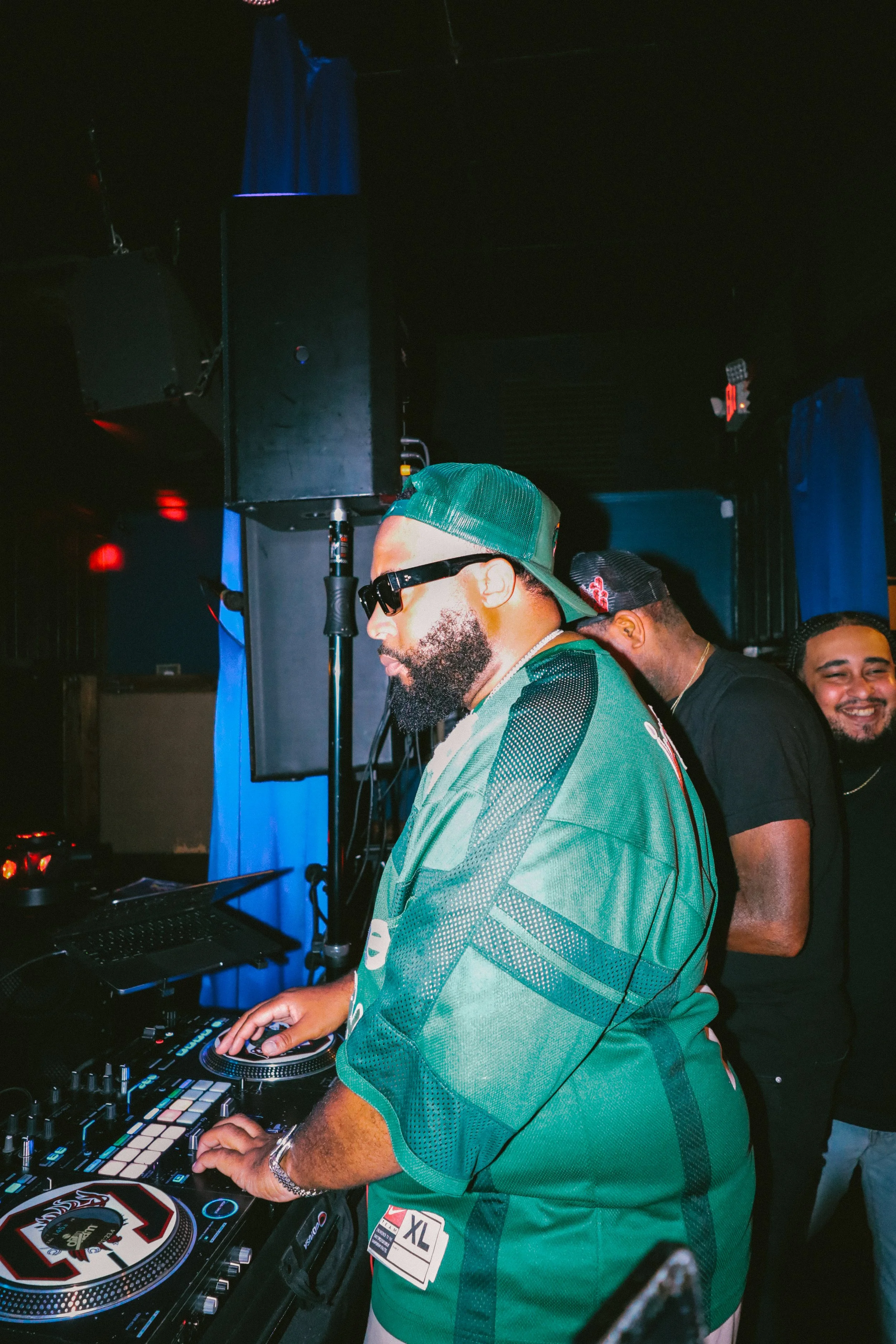 A man wearing a green jersey, black sunglasses, and a green cap DJing on a mixing console at a nightclub. There are people standing behind him, and the setting appears dark with some blue curtains and large speakers in the background.