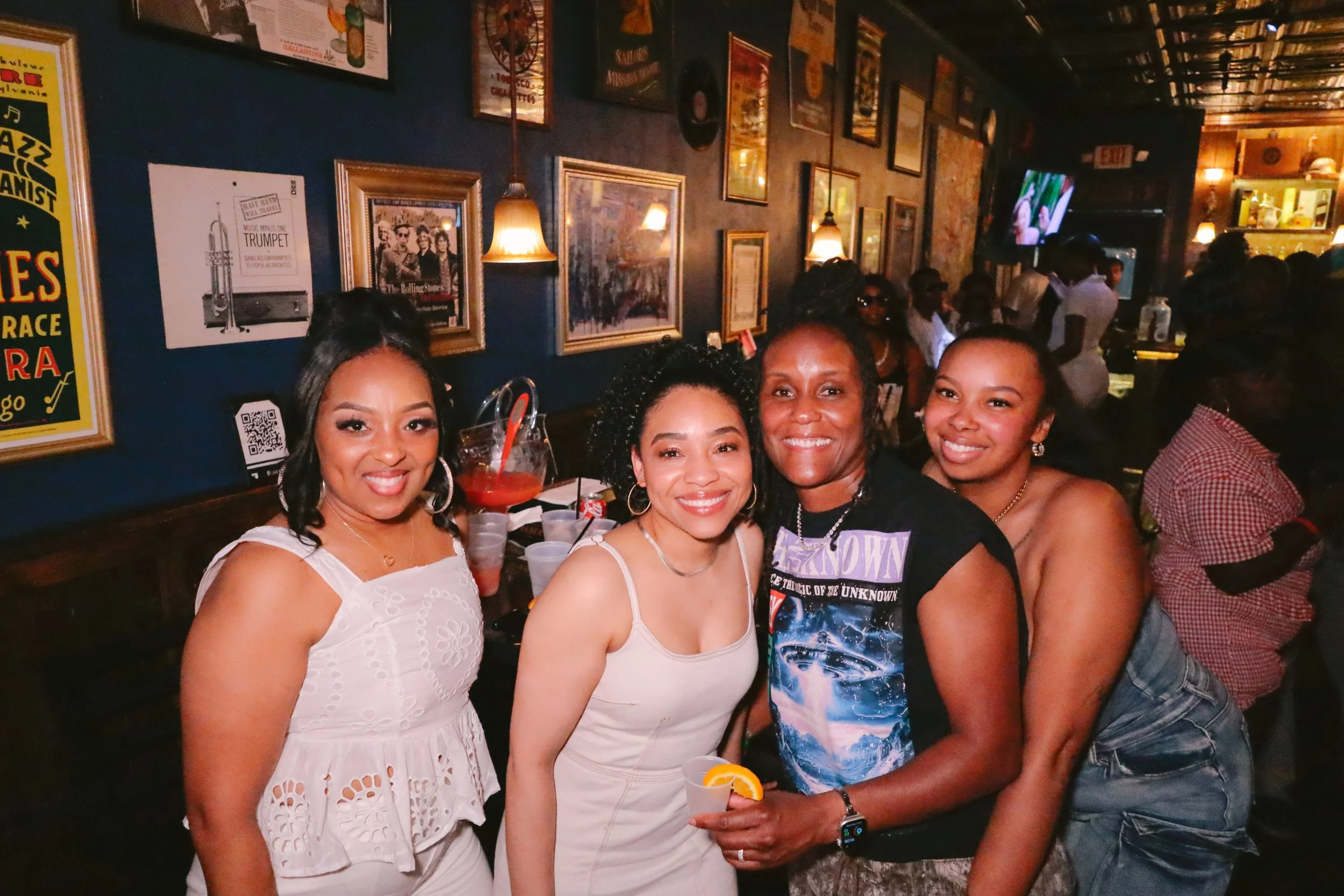 Four women smiling and posing for a photo at a lively indoor party or bar with framed pictures and posters on the wall behind them, some holding drinks.