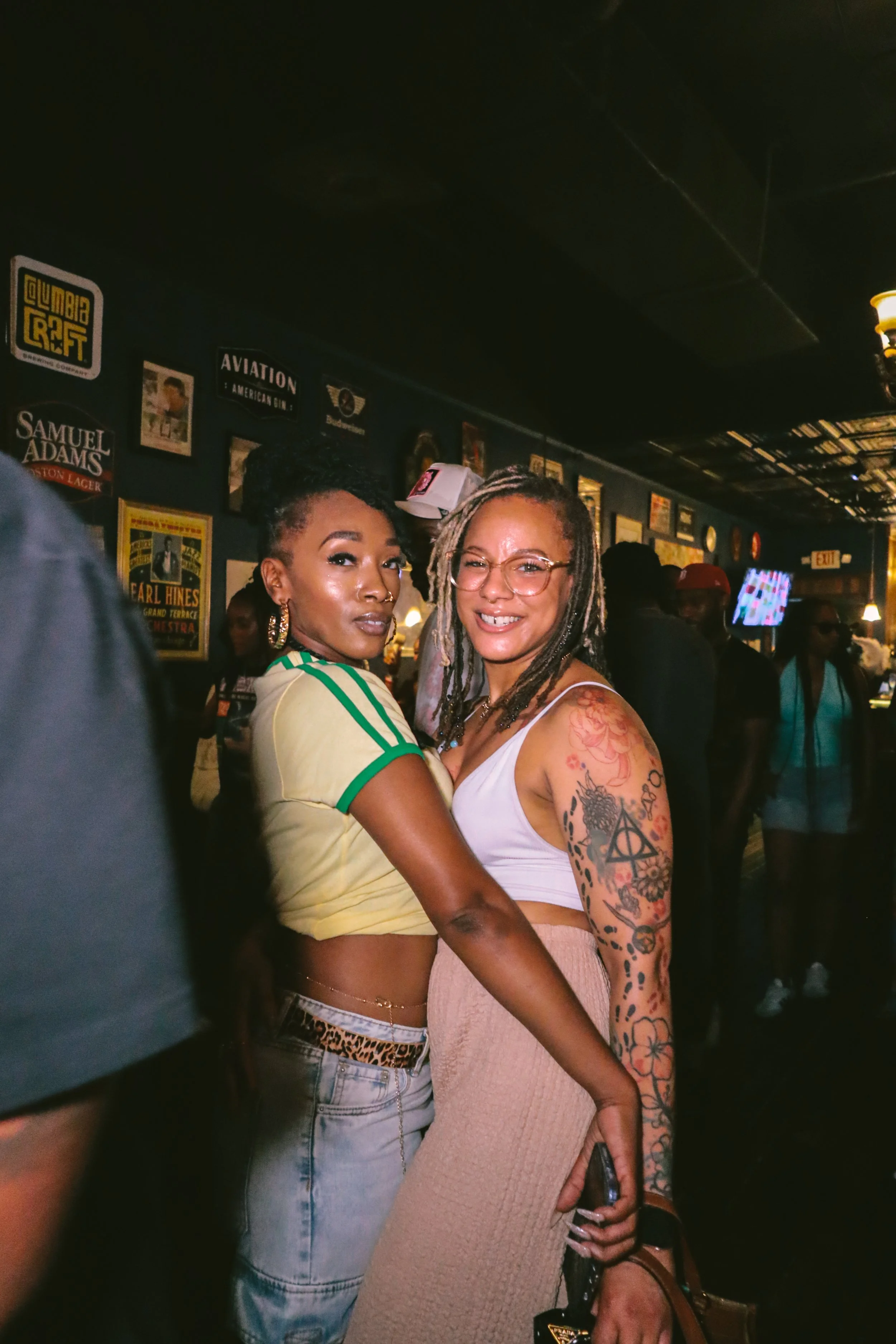 Two women pose together in a bar or club, with one wearing a yellow athletic top and the other a white tank top with tattoos on her arms, surrounded by people and dim lighting.