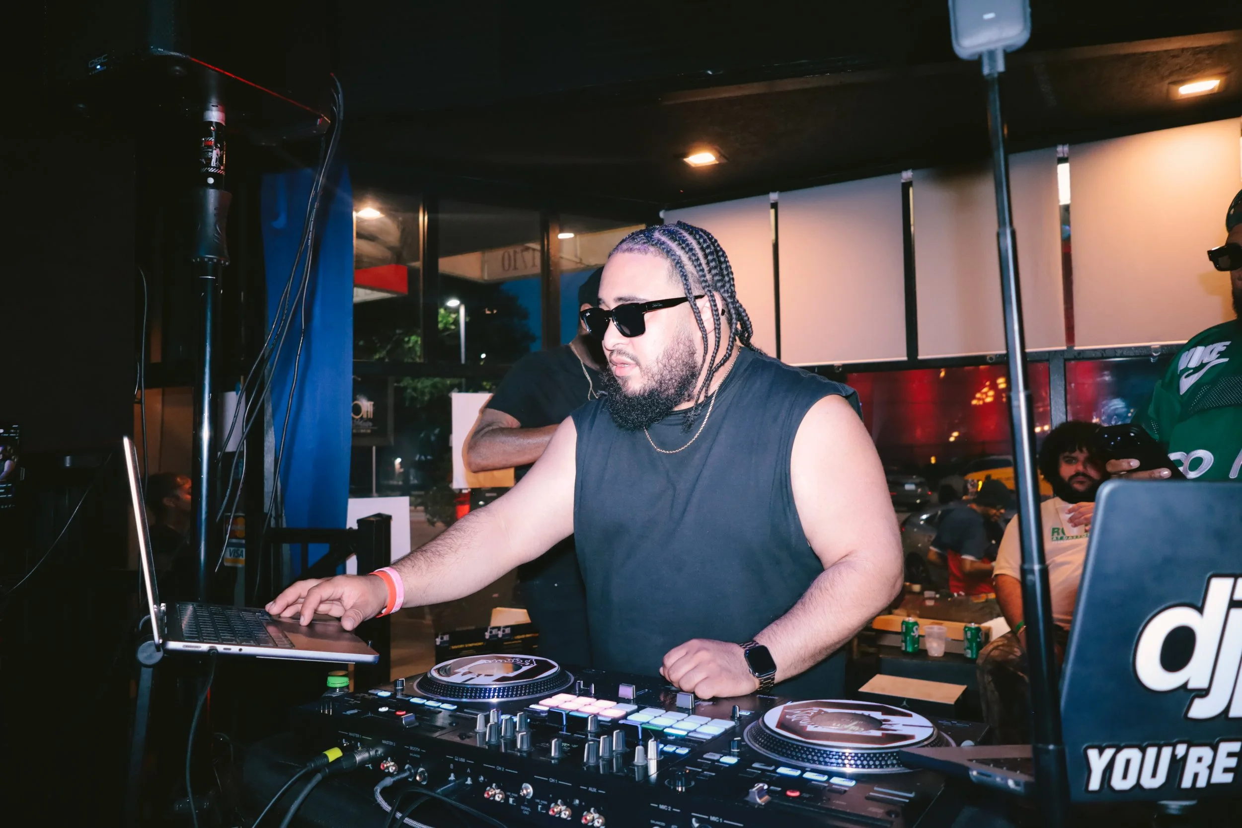 A DJ with braided hair, sunglasses, and a black sleeveless shirt is performing at an indoor event with people in the background. DJ equipment, including a laptop and turntables, is in front of him.