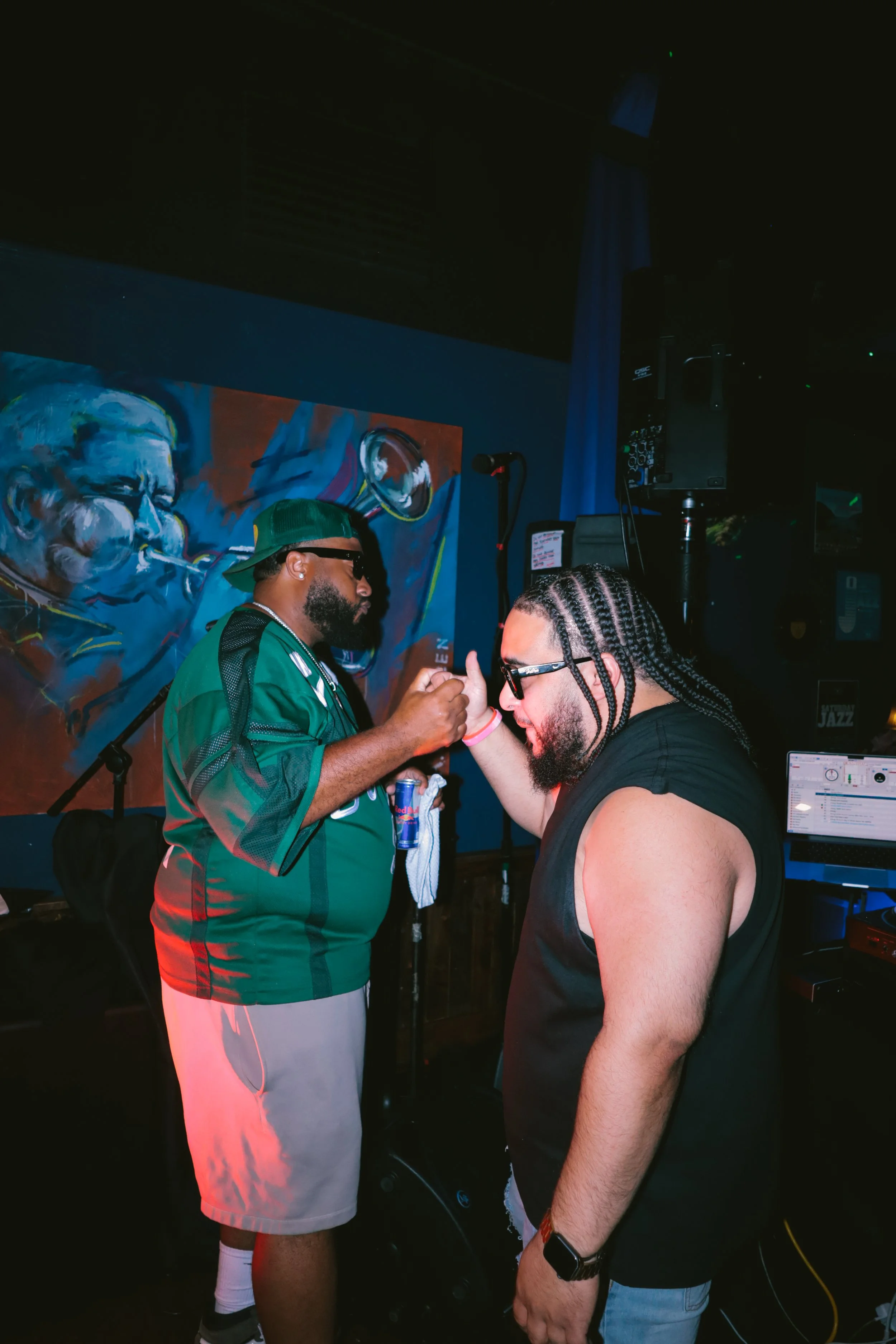 Two men are dancing and holding hands or arms in a nightclub or music venue. One man has long braided hair, glasses, and a black sleeveless shirt, while the other man has a beard, sunglasses, and is wearing sneakers, shorts, and a cap. There is a col