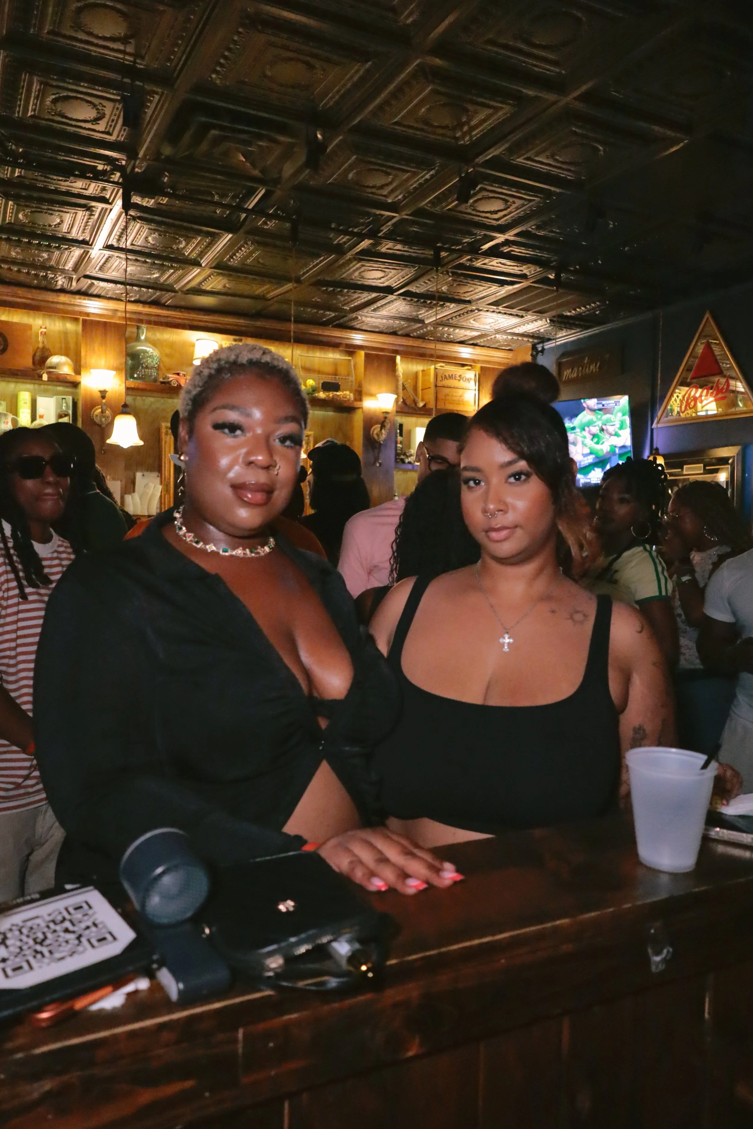 Two women sitting at a bar or restaurant, with others in the background. One woman has short, blonde hair, dark lipstick, and is wearing a black top with a colorful necklace. The other woman has shoulder-length brown hair, tattoos on her shoulder and