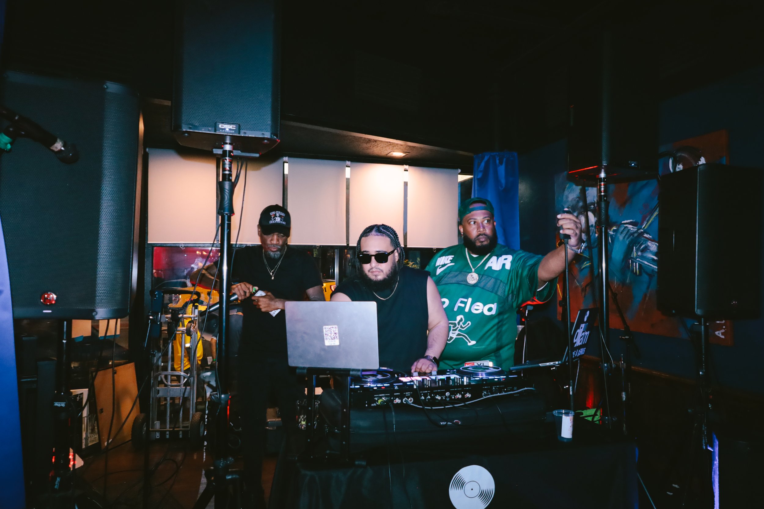 Three men are behind DJ equipment and speakers in a dimly lit indoor space with dark walls and colorful artwork. One man in sunglasses is centered, another man with a beard and cap to the right, and a third man in a black cap to the left.