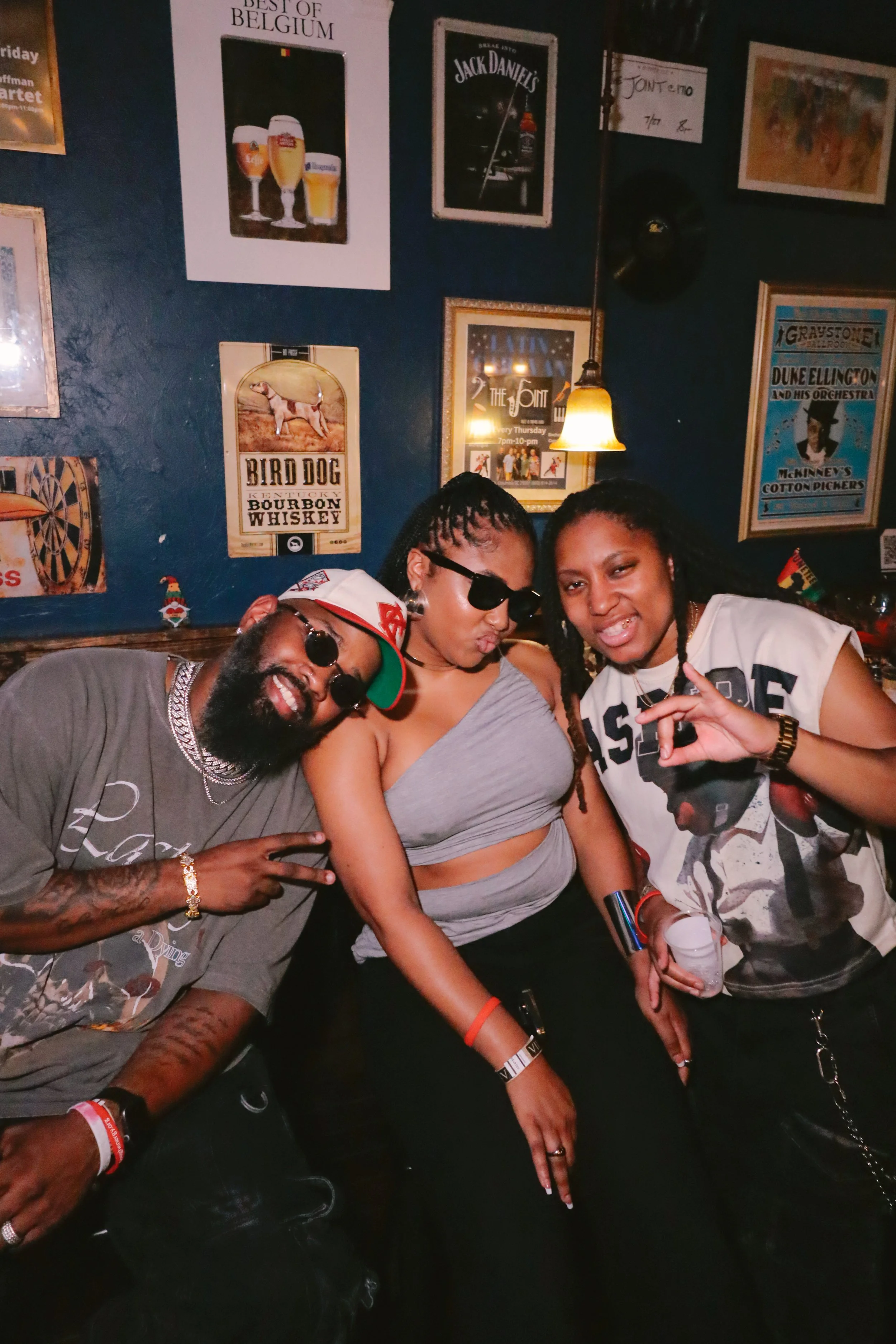 Three young adults in a bar or nightclub, posing together and making hand signs. The background features framed posters related to beverages and music.