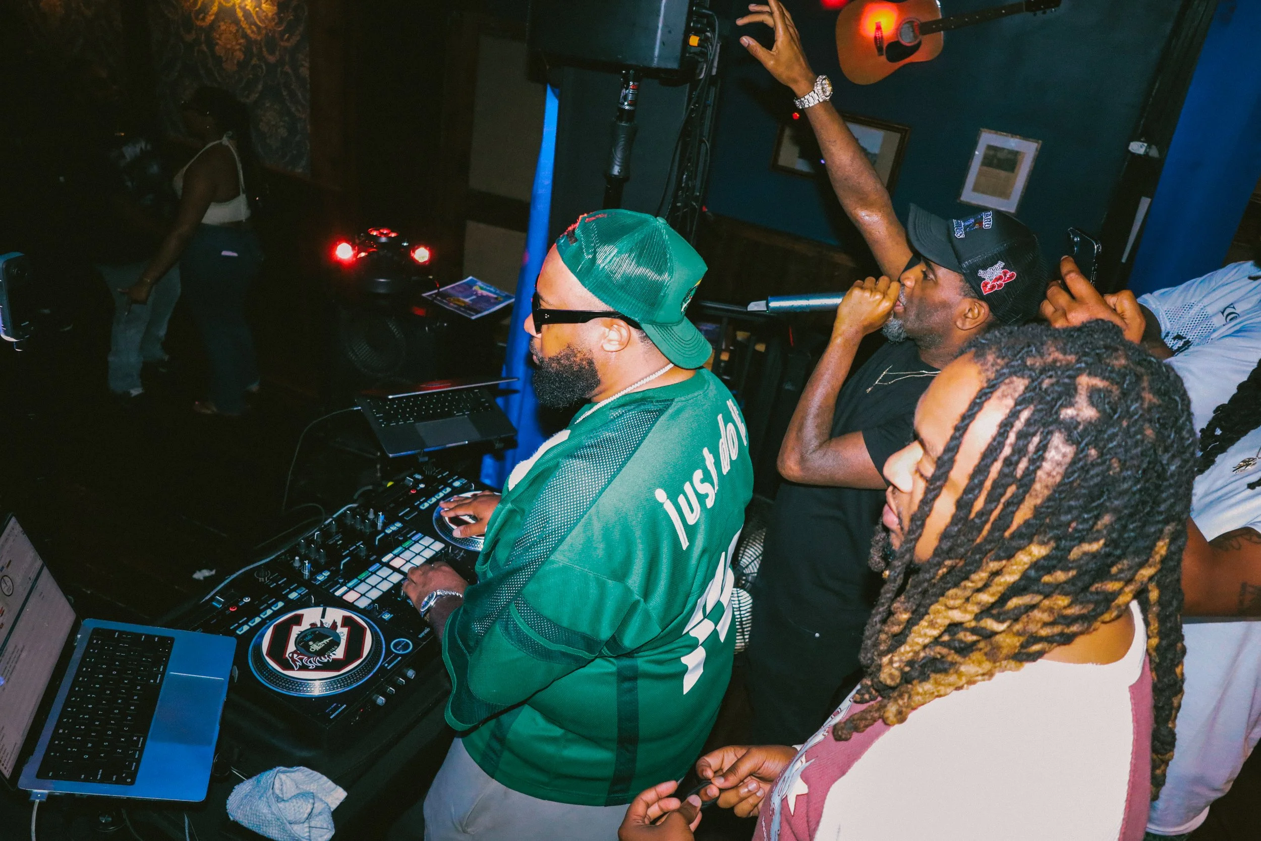 DJ iAM wearing a green cap and sunglasses operates a DJ controller in a dimly lit room, with a man in a black cap and shirt leaning in, and a woman with braided hair observing nearby.
